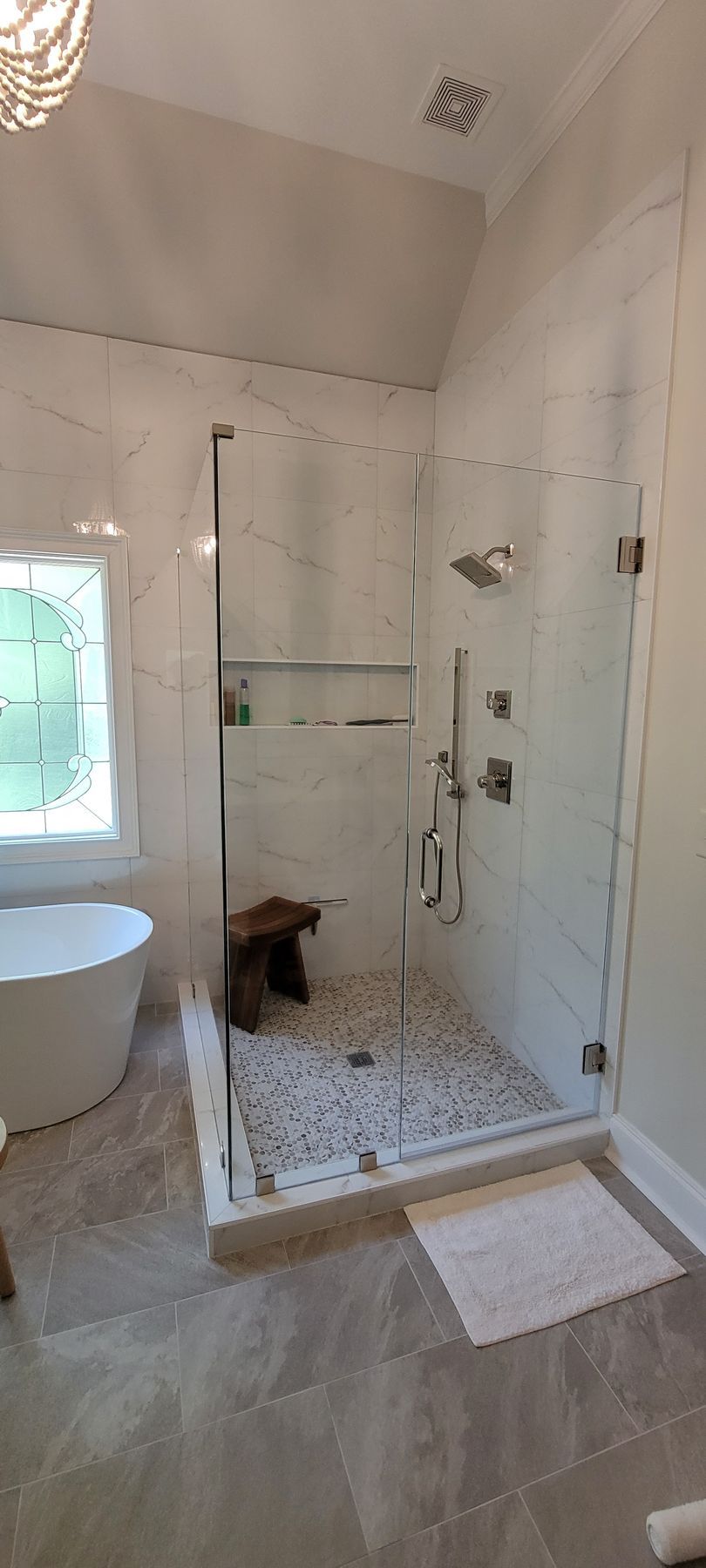Bathroom with walk-in glass shower, white tile, and beige floor; window beside a tub and rug in front of shower.