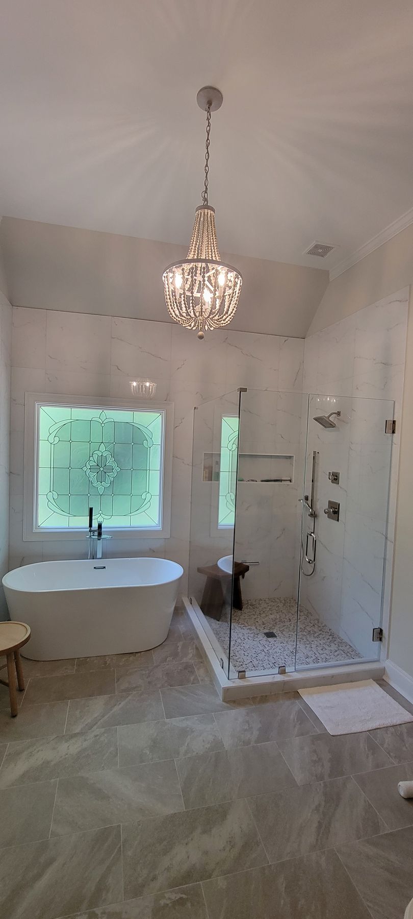 Bright white bathroom with chandelier, freestanding tub, glass shower, and two frosted windows