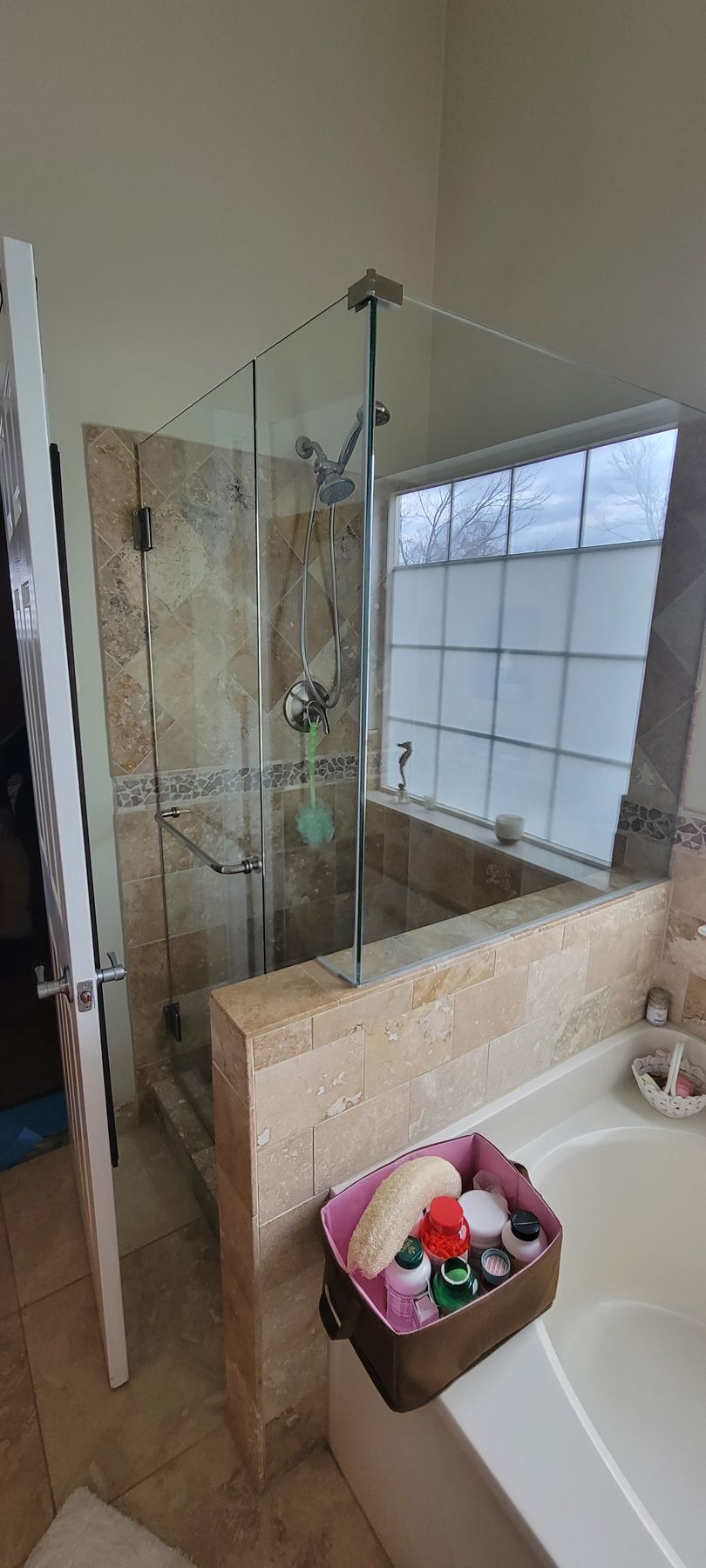 Bathroom with a glass shower enclosure, stone wall, window, and a basket of toiletries by the tub