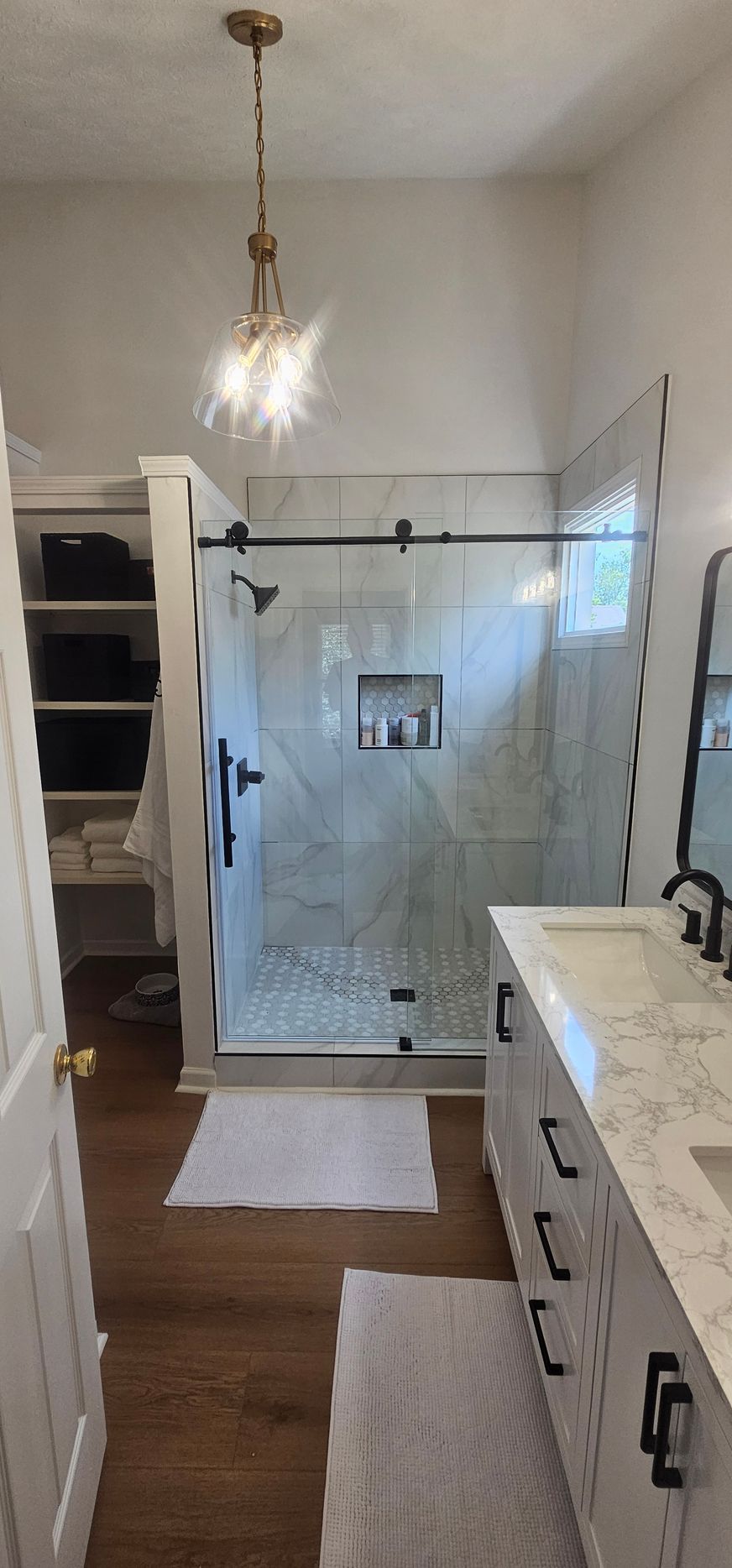 Bathroom with glass shower enclosure, vanity, shelving, and a hanging light over a wood floor.