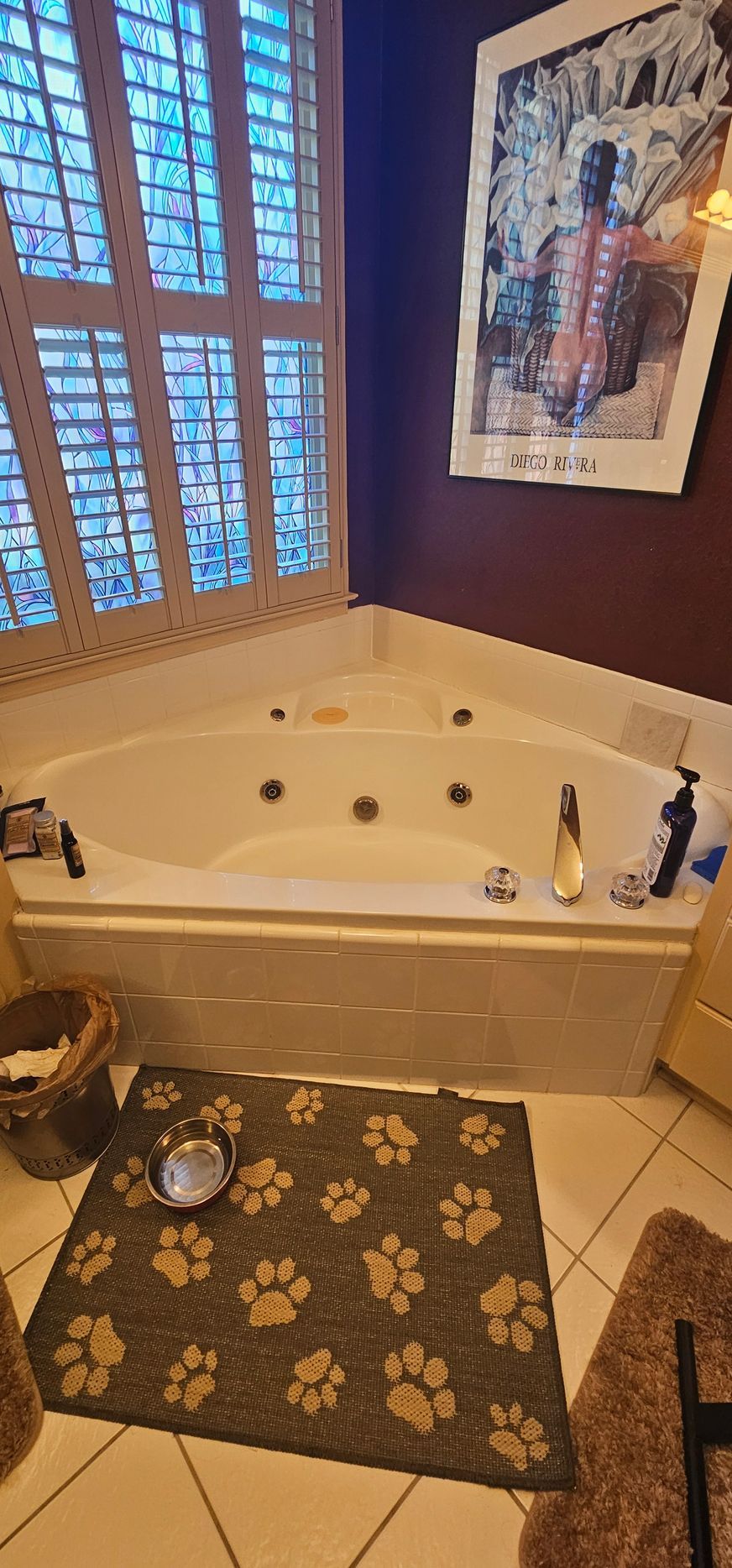 Corner bathroom with a white corner tub, brown walls, floral rug, and window with plantation shutters.