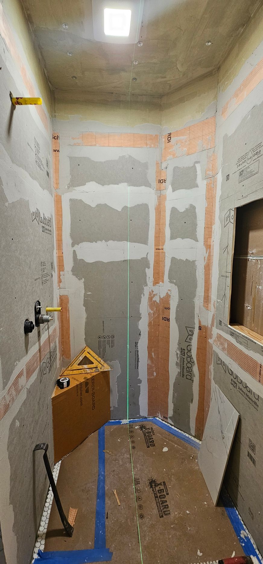 Narrow unfinished room with drywall patching, ladder, and blue painter’s tape on the floor.