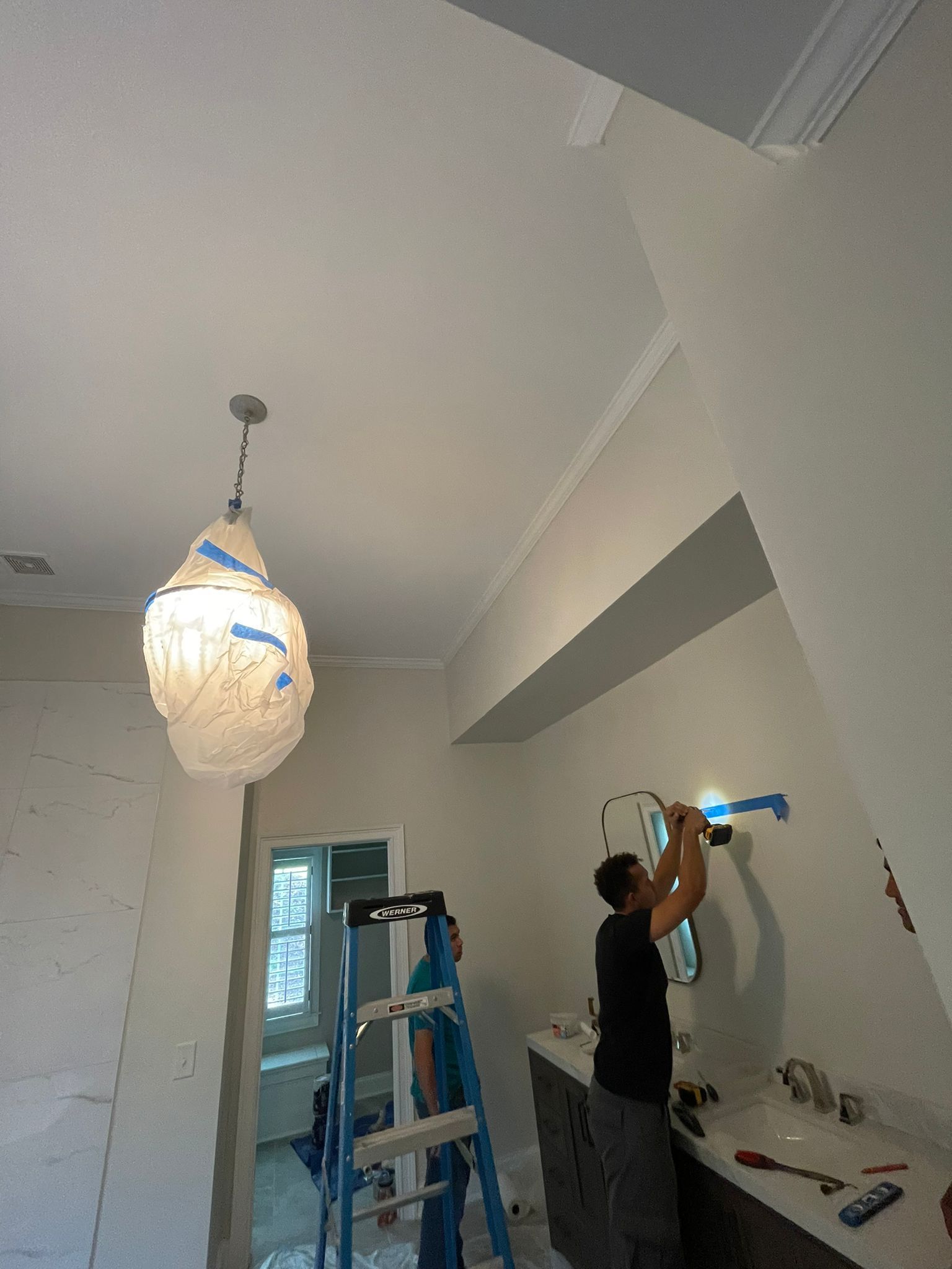 Person working on a wall inside a room, using a blue hand tool near a ladder and hanging light fixture.