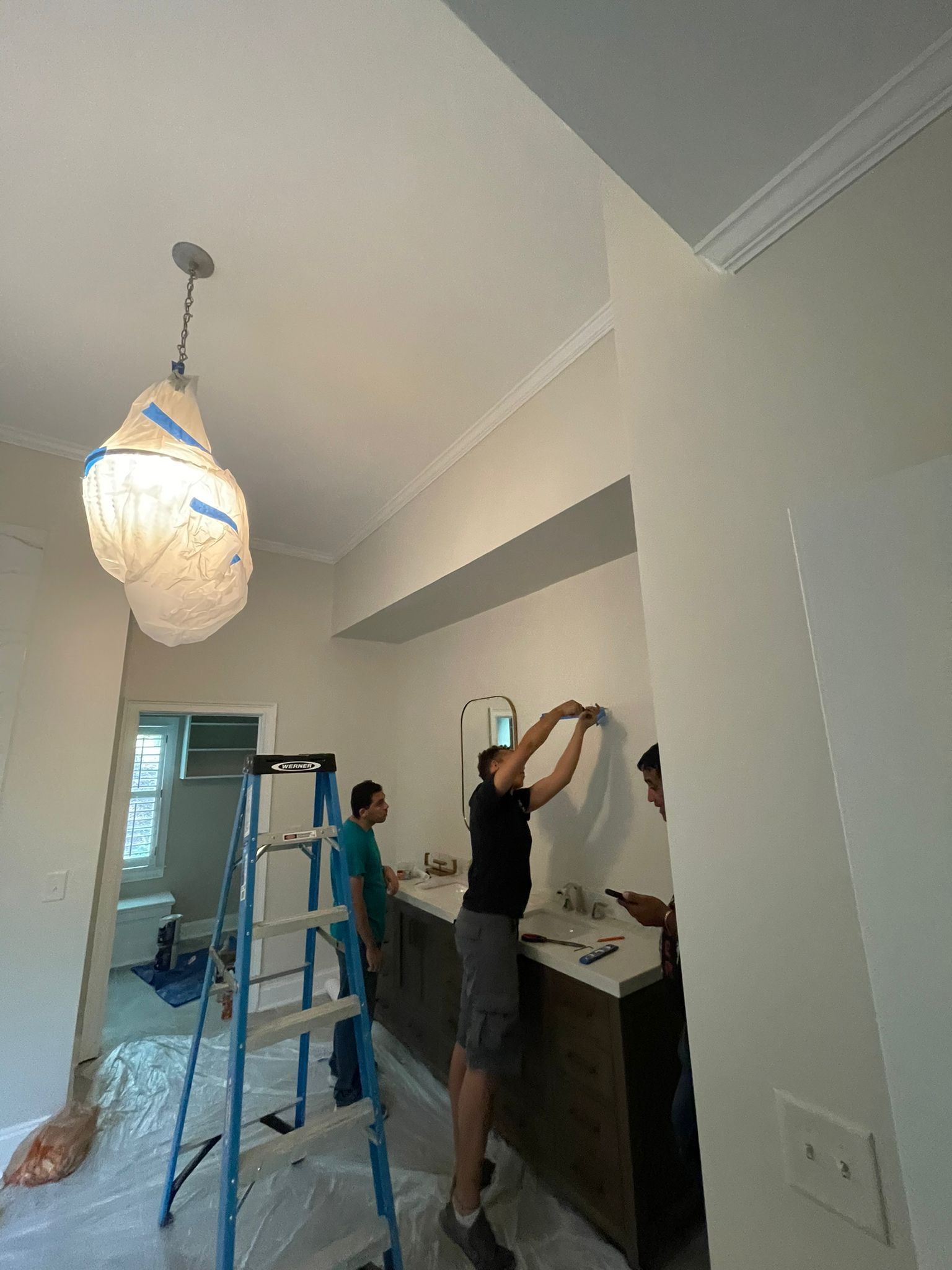 Person painting a hallway wall beside a ladder and cabinet under a hanging light fixture