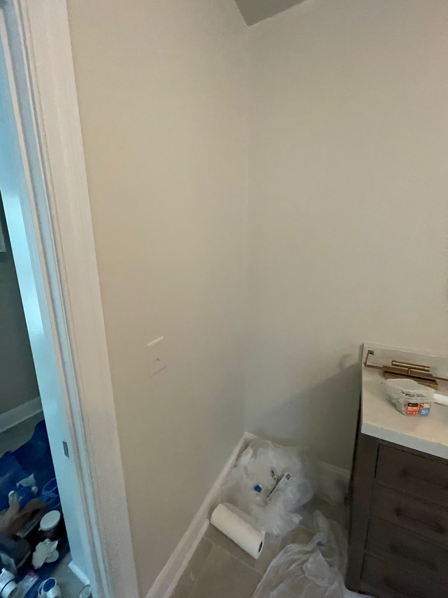 Small room corner with paint supplies on the floor beside a dresser and open doorway