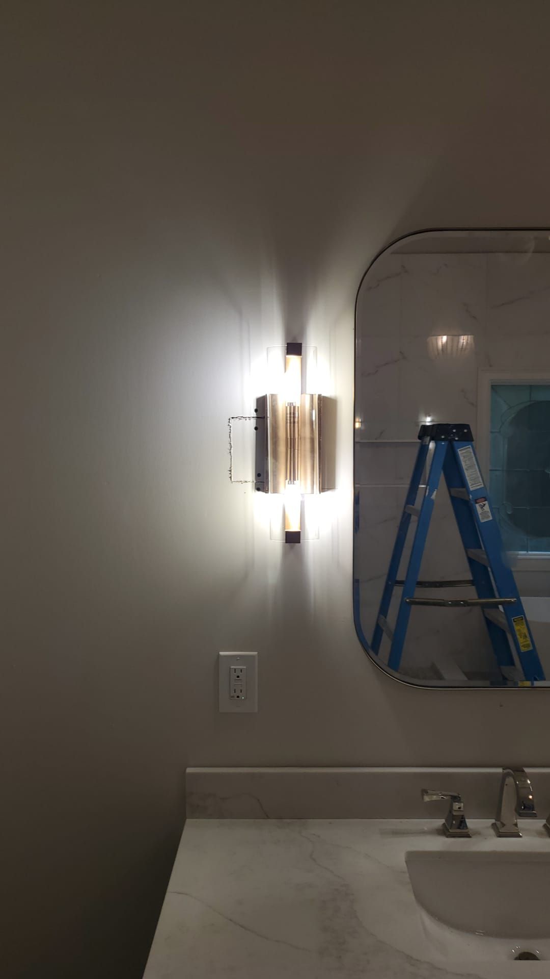 Bathroom vanity with lit wall sconces, mirror, sink, and a blue step ladder reflected in the mirror.