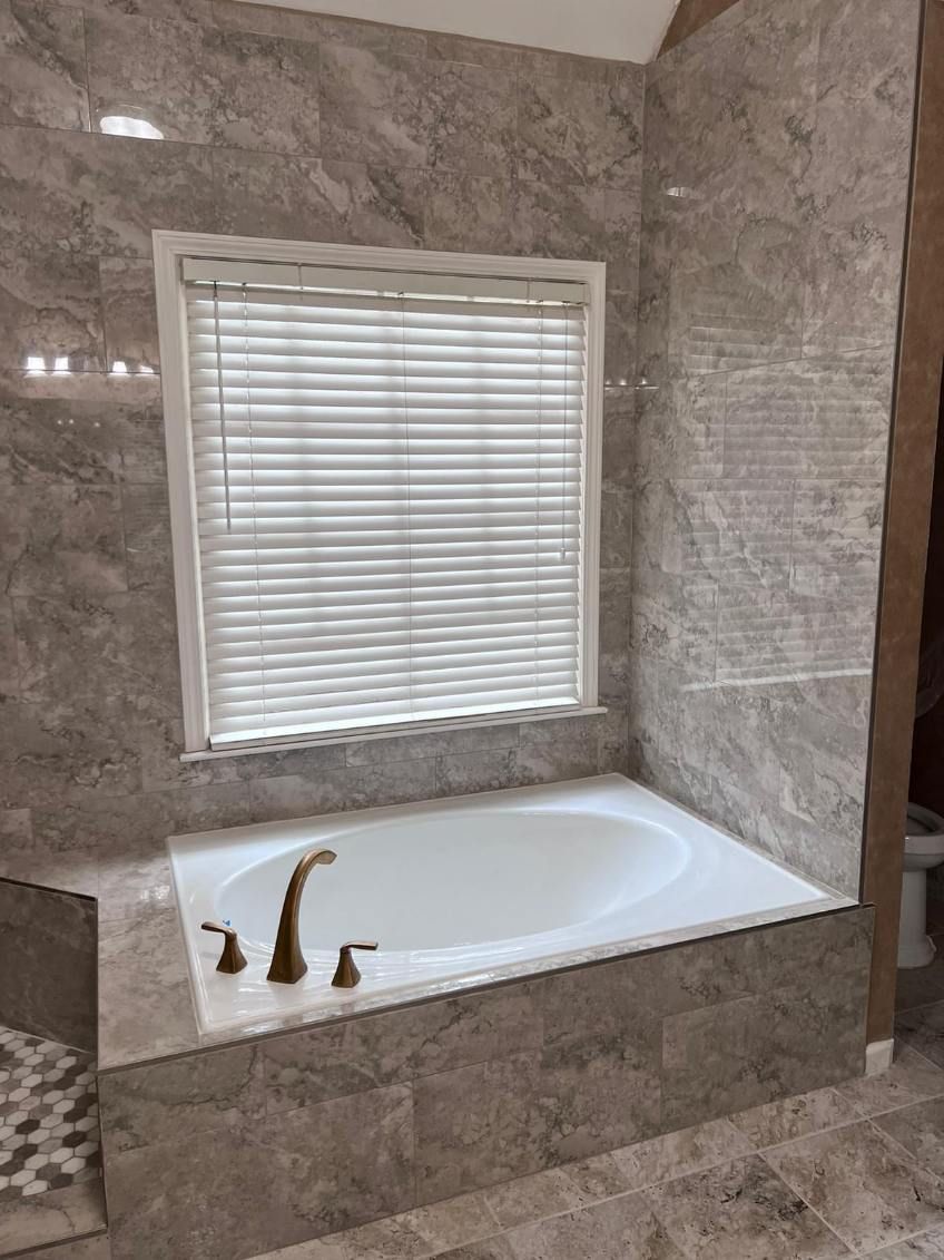 Bathroom with tiled tub alcove, white bathtub, and window with blinds.