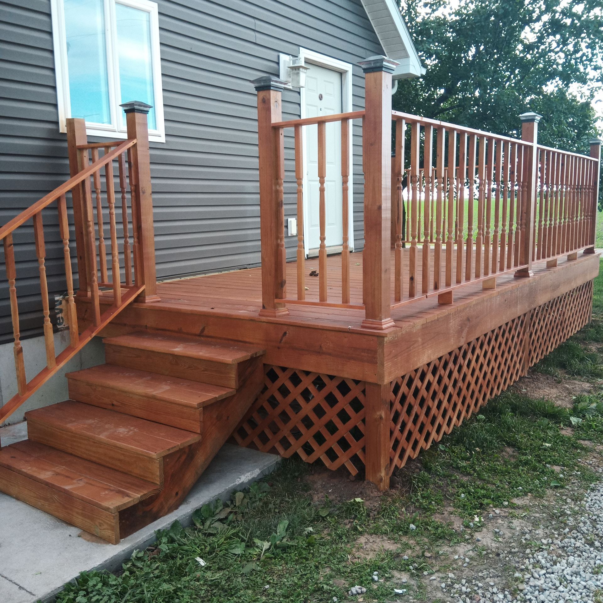 Wooden deck with stairs, railing, and lattice skirting attached to a gray house.