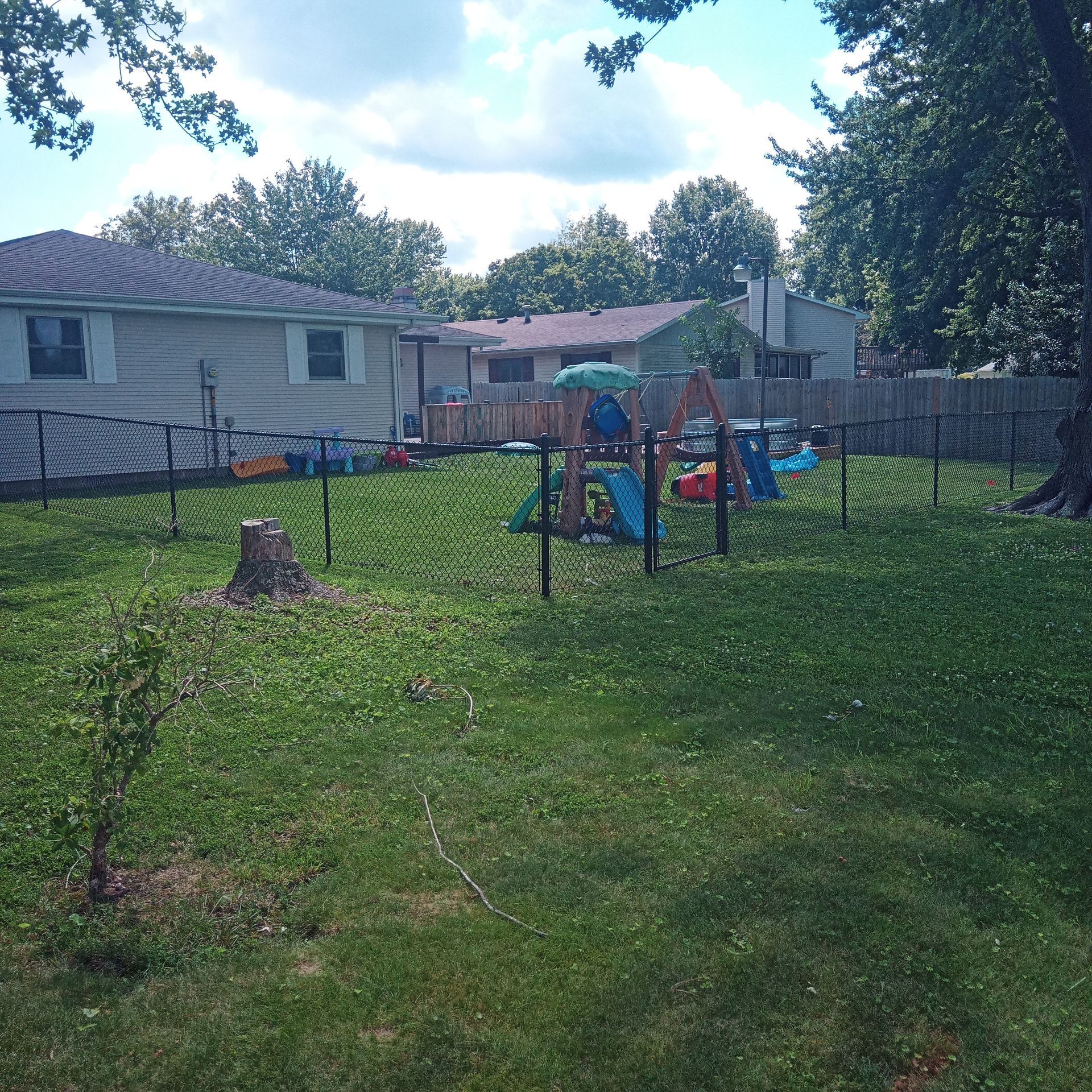 Backyard with a playground, enclosed by a black fence. Houses and trees are in the background, under a partly cloudy sky.