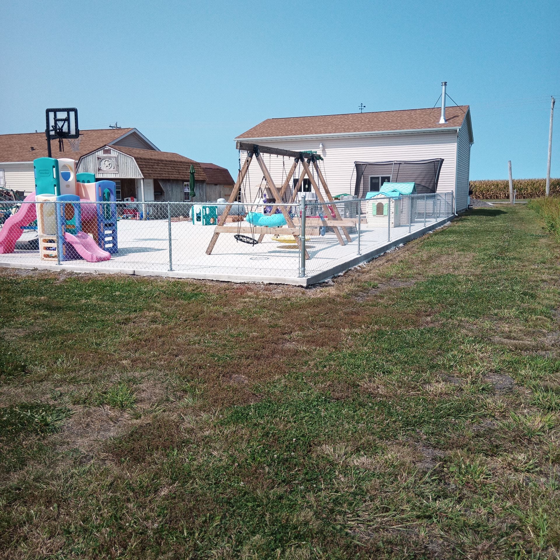 Playground with swings, slides, and a basketball hoop, enclosed by a chain-link fence, under a blue sky.