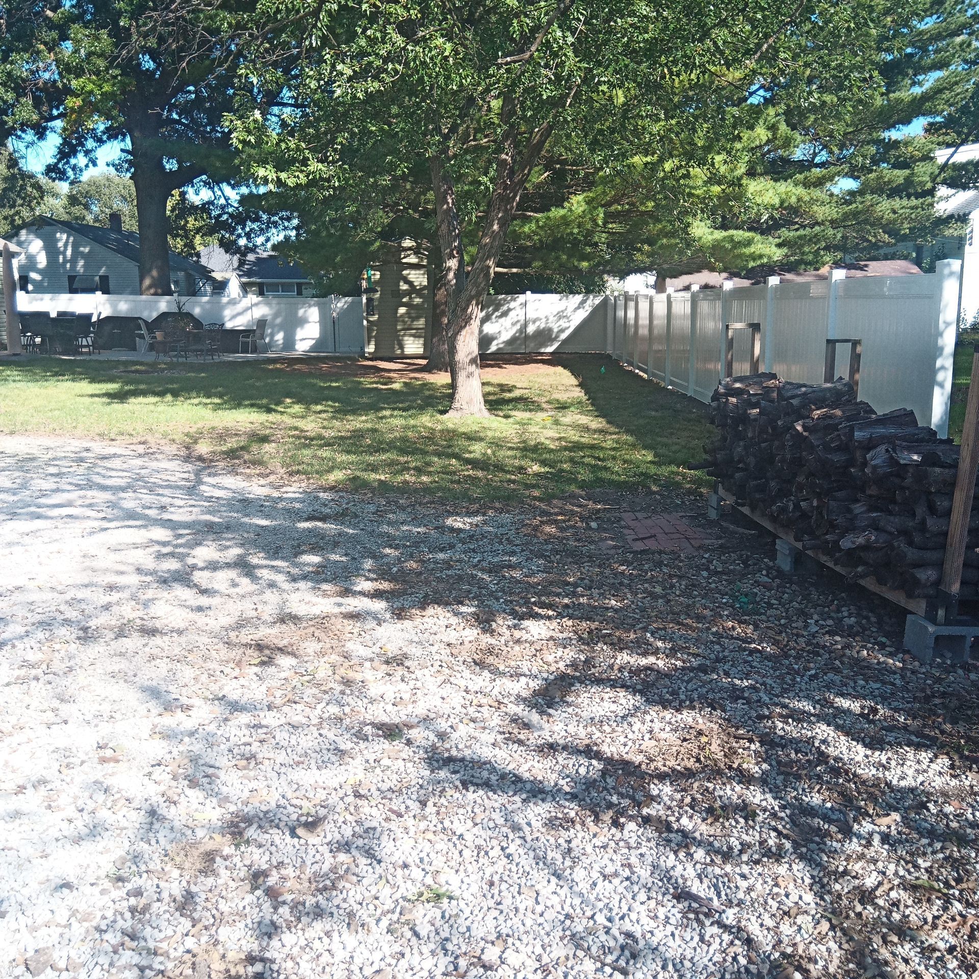 Gravel area with grass and trees next to a white fence. Firewood is stacked along the fence.