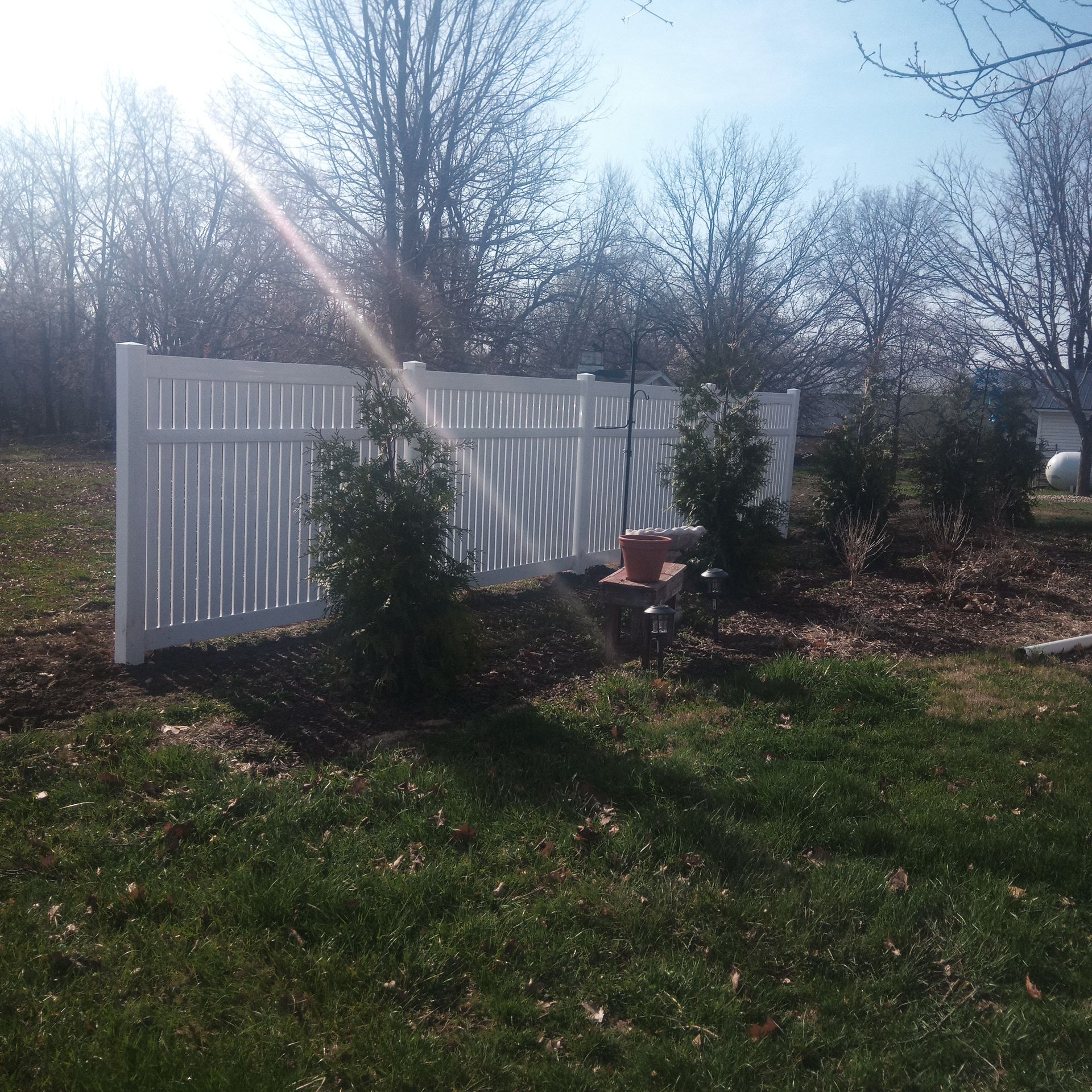 White vinyl fence in a grassy yard, sunlight shining, trees in the background.