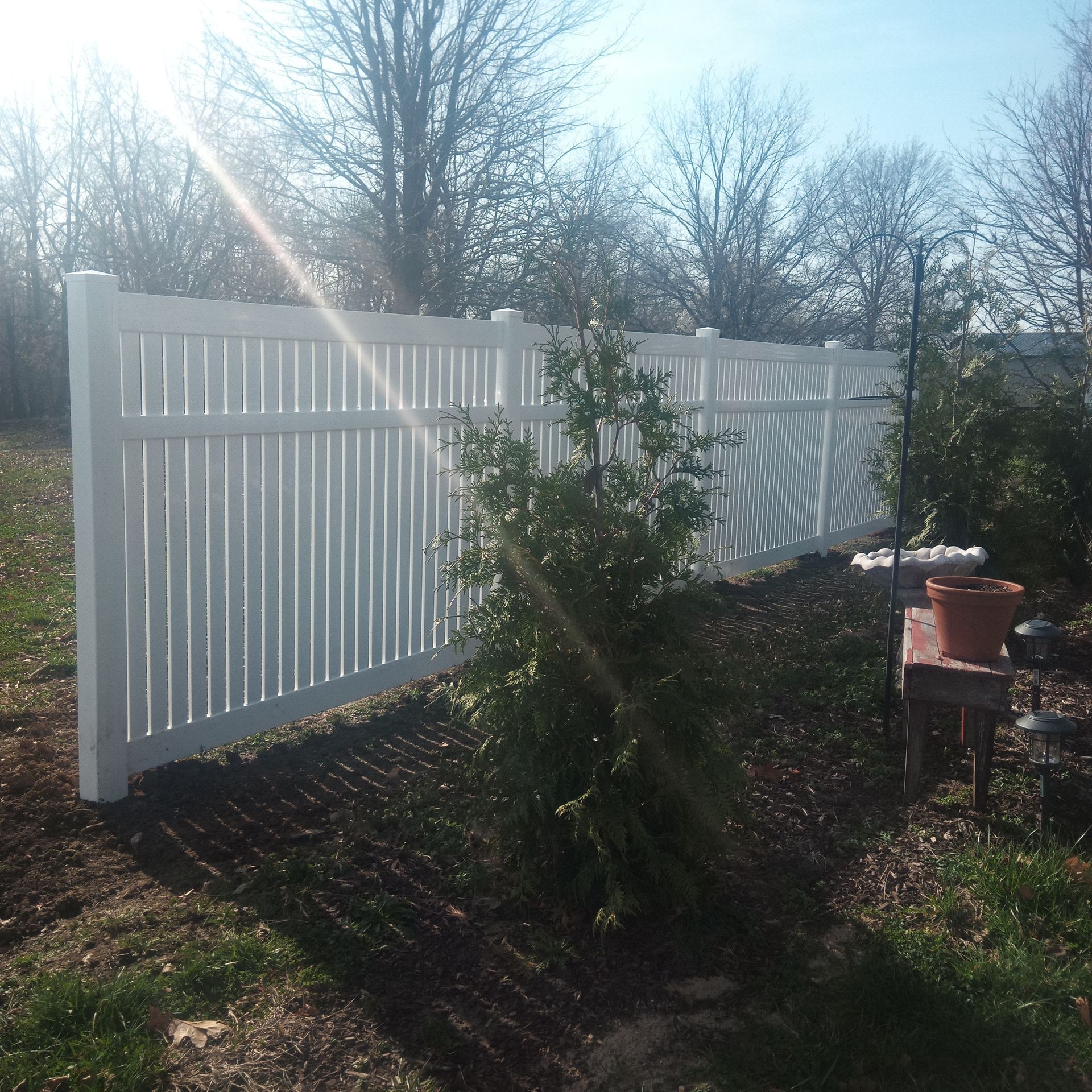 White vinyl fence in a sunny backyard, with a small tree and potted plant visible.