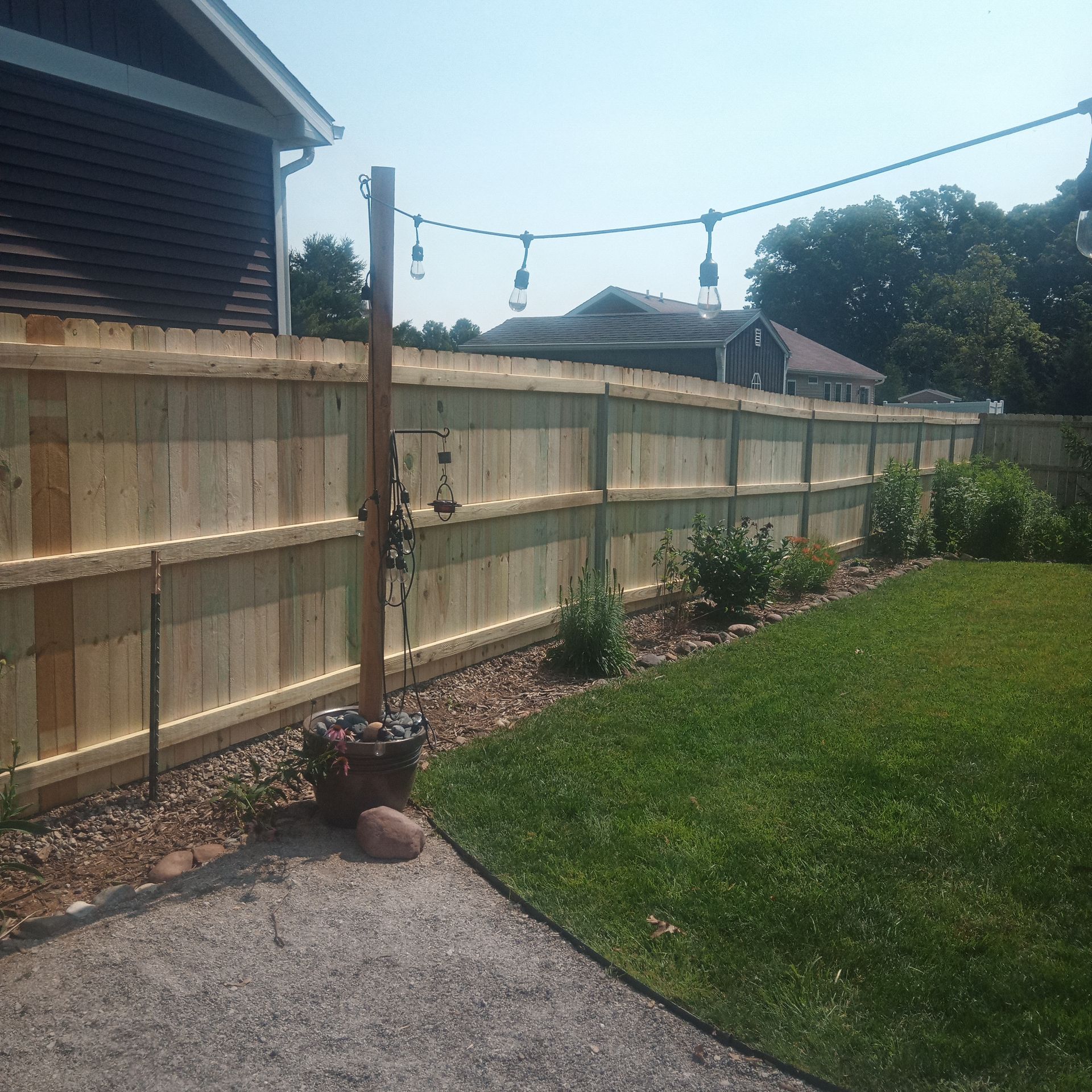 Wooden fence in a backyard with string lights overhead and grass on the right.