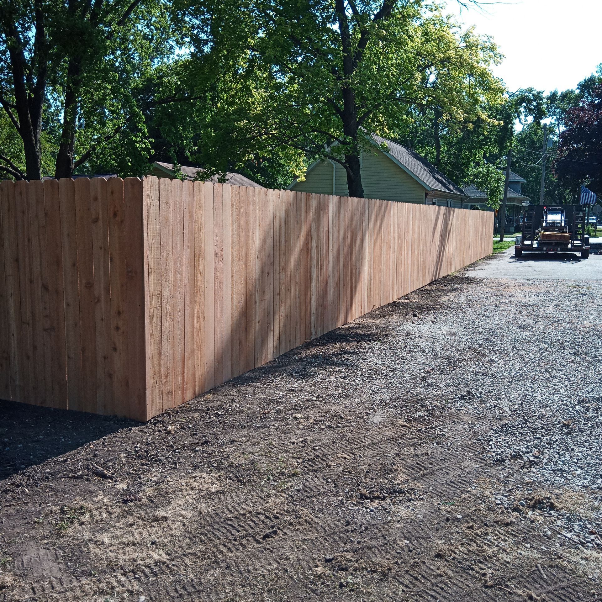 Wooden fence along a gravel driveway; trees in the background, a vehicle is parked nearby.