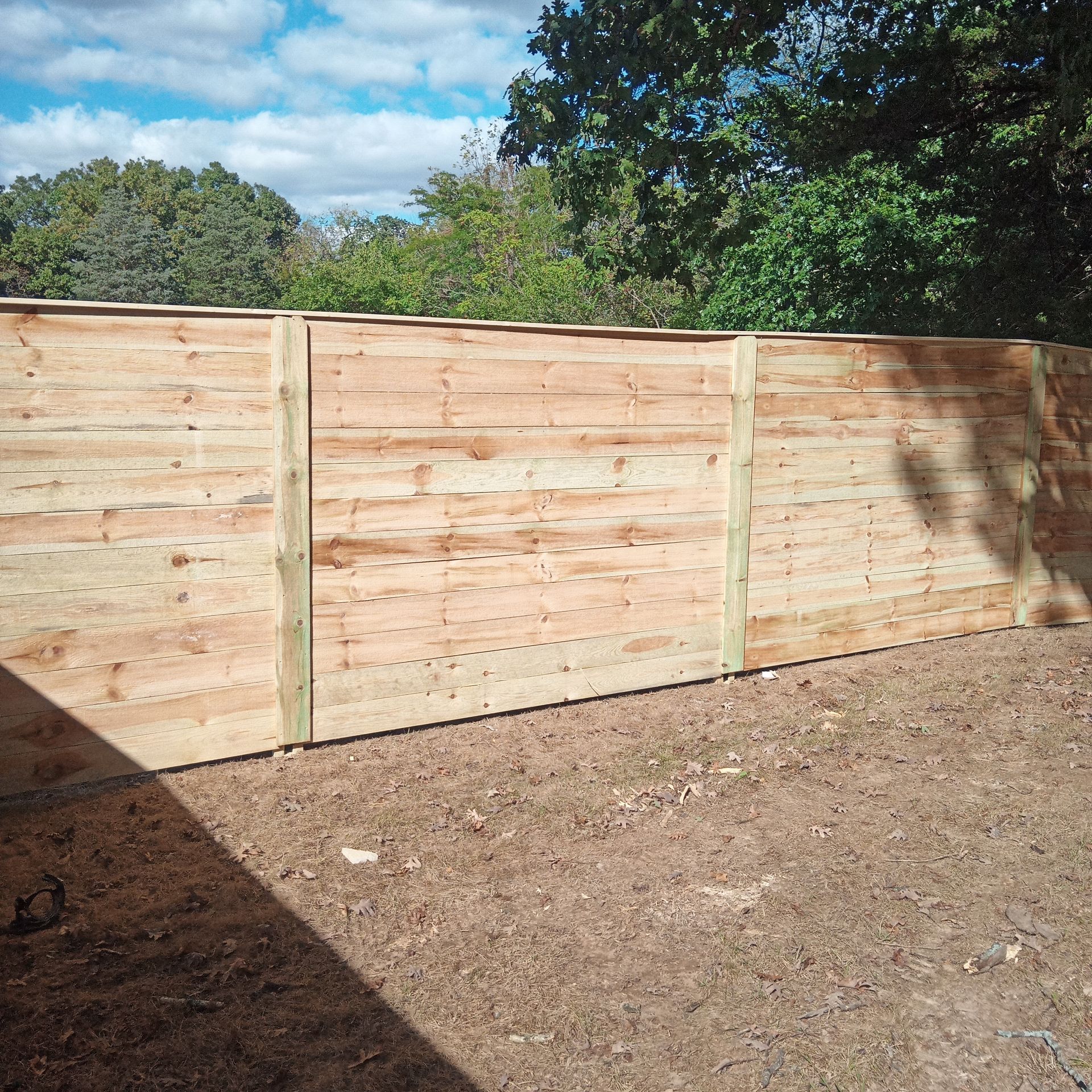 Wooden fence in front of trees, with posts, and a dirt ground.