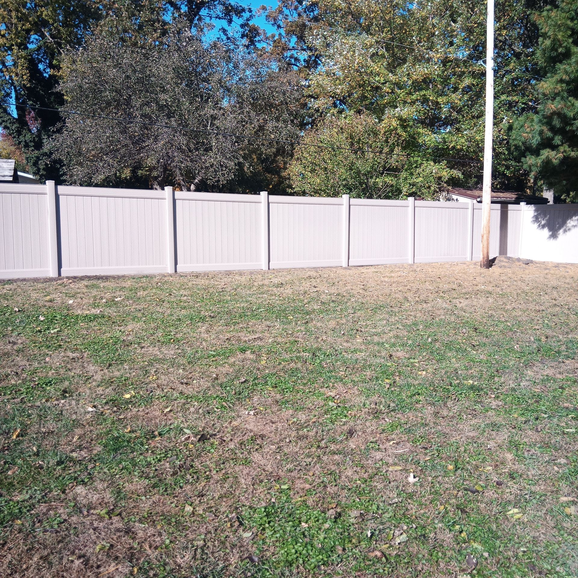 Tan vinyl fence enclosing a grassy yard, trees in background, clear sky.