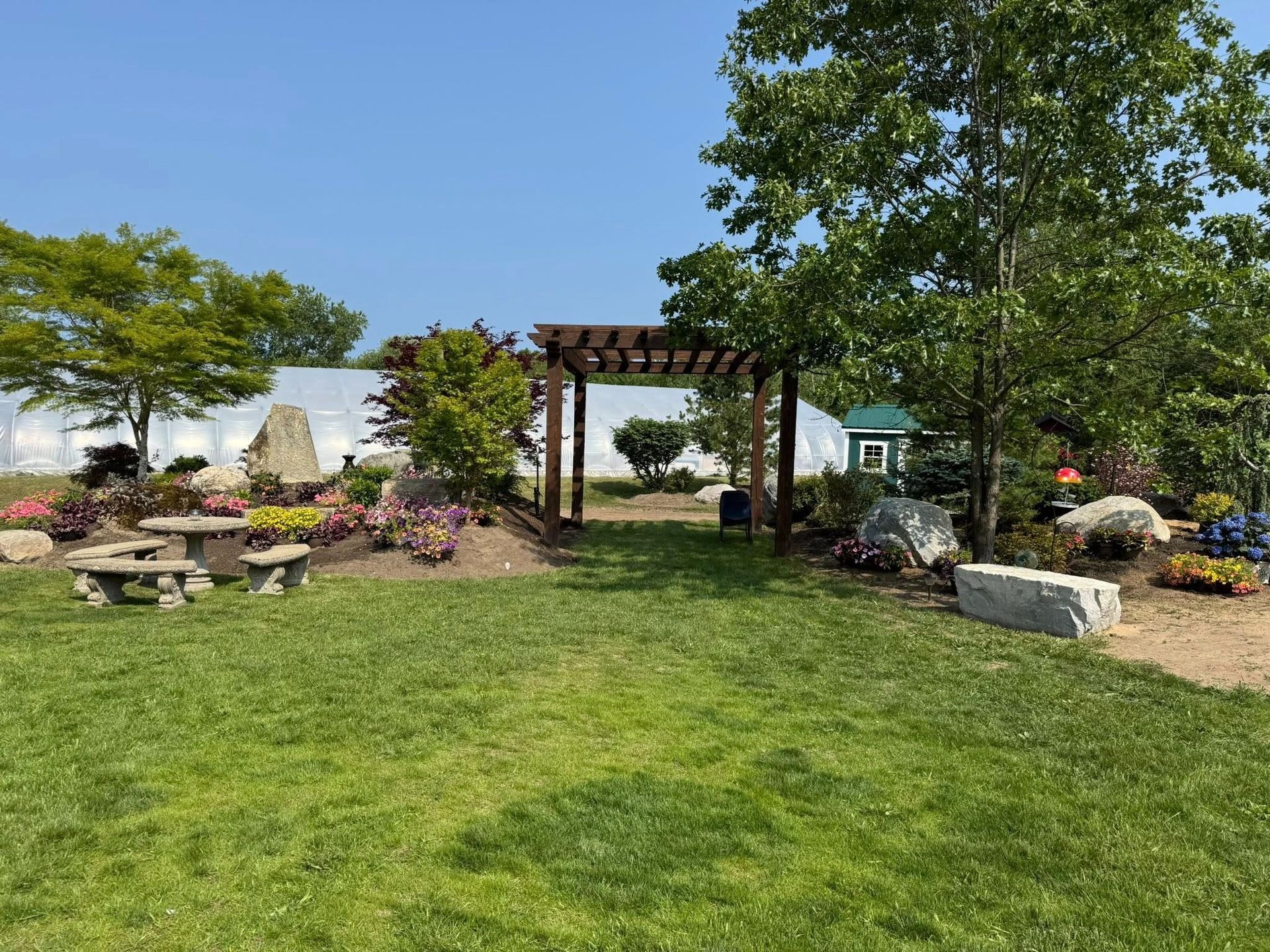 A grassy garden area with a wooden pergola, flower beds, and picnic tables under a sunny sky.