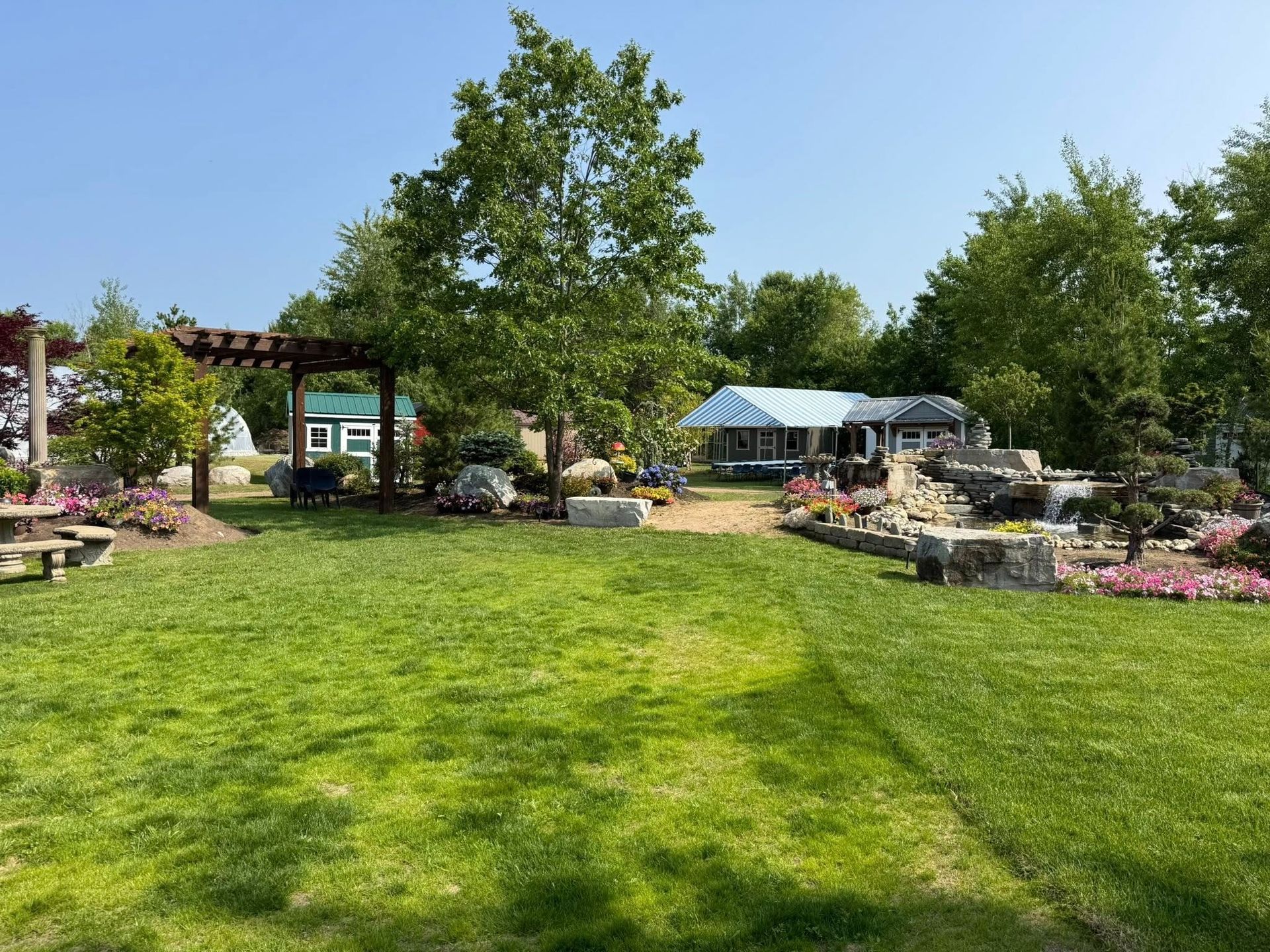 Green grassy park with pergola, tree, and a small building with blue roof.