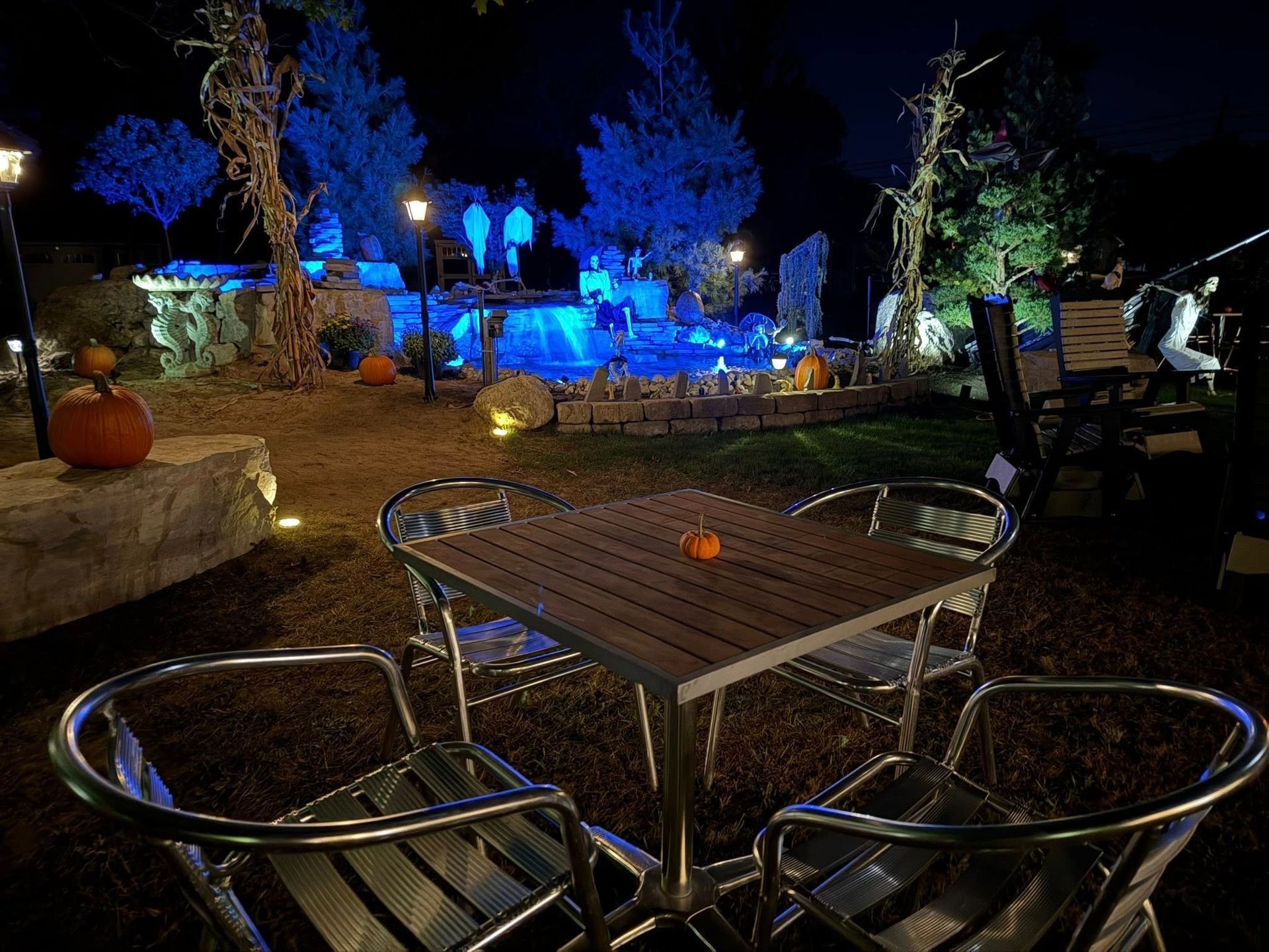 Outdoor Halloween scene with tables and chairs, illuminated by blue lights, with pumpkins and water feature in background.