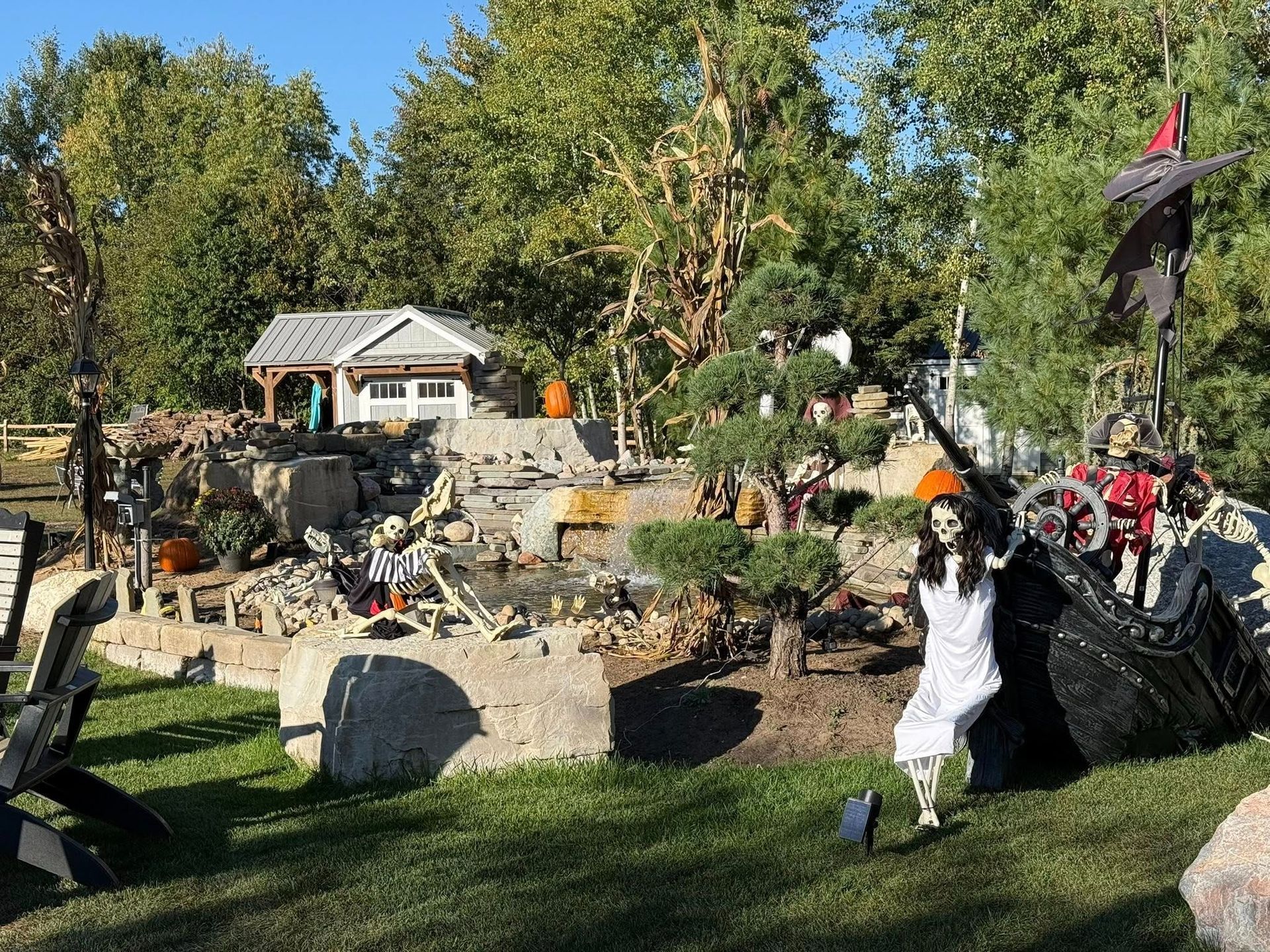 Halloween yard display with skeletons, pirate ship, pond, and small white house.