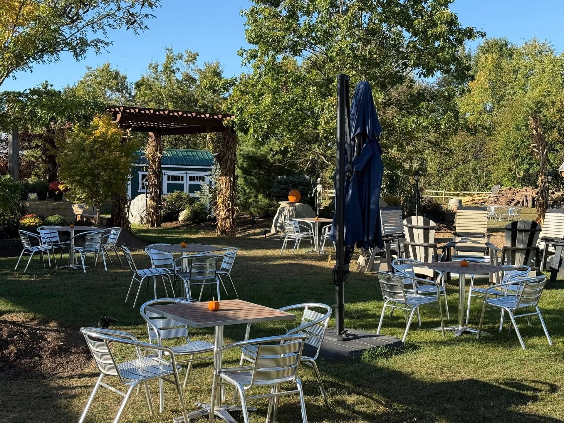 Outdoor seating area with tables and chairs on a grassy lawn; a blue umbrella is closed, and trees surround the area.