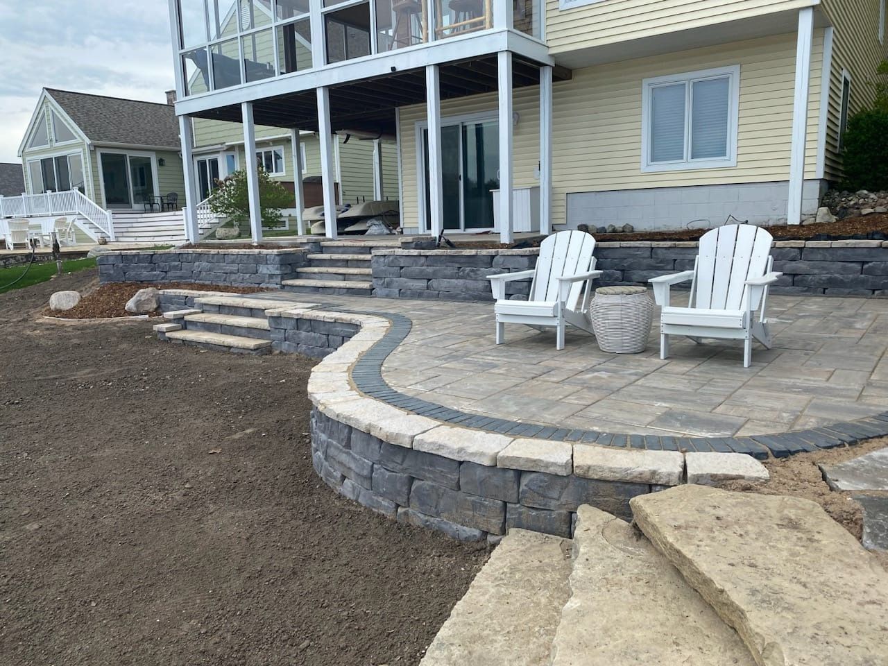 Stone patio with two white chairs, next to a two-story yellow house with a glassed-in balcony.