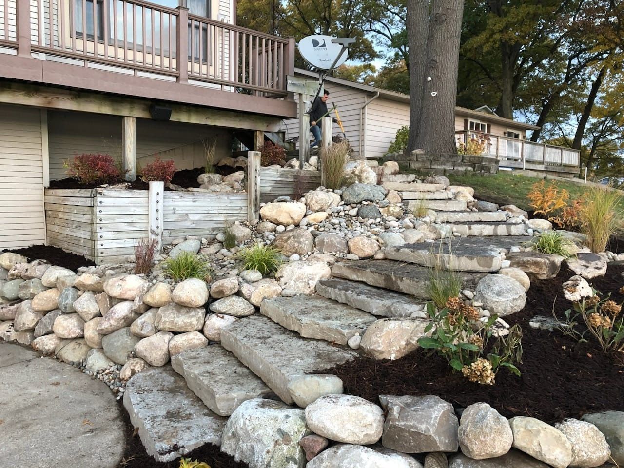 Stone steps and landscaping leading up to a lakeside home; a person stands near the top.