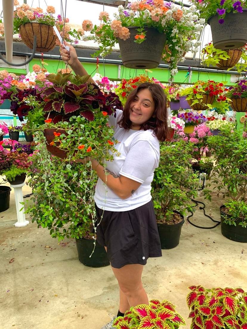 Woman in a greenhouse holding a hanging basket of colorful flowers, smiling. Other flower baskets are in the background.