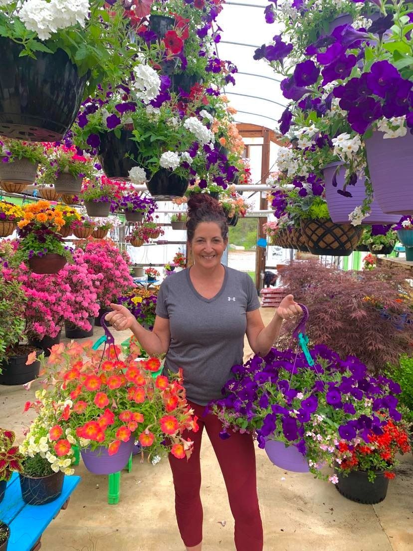 Woman holding two hanging flower baskets among many vibrant hanging plants.