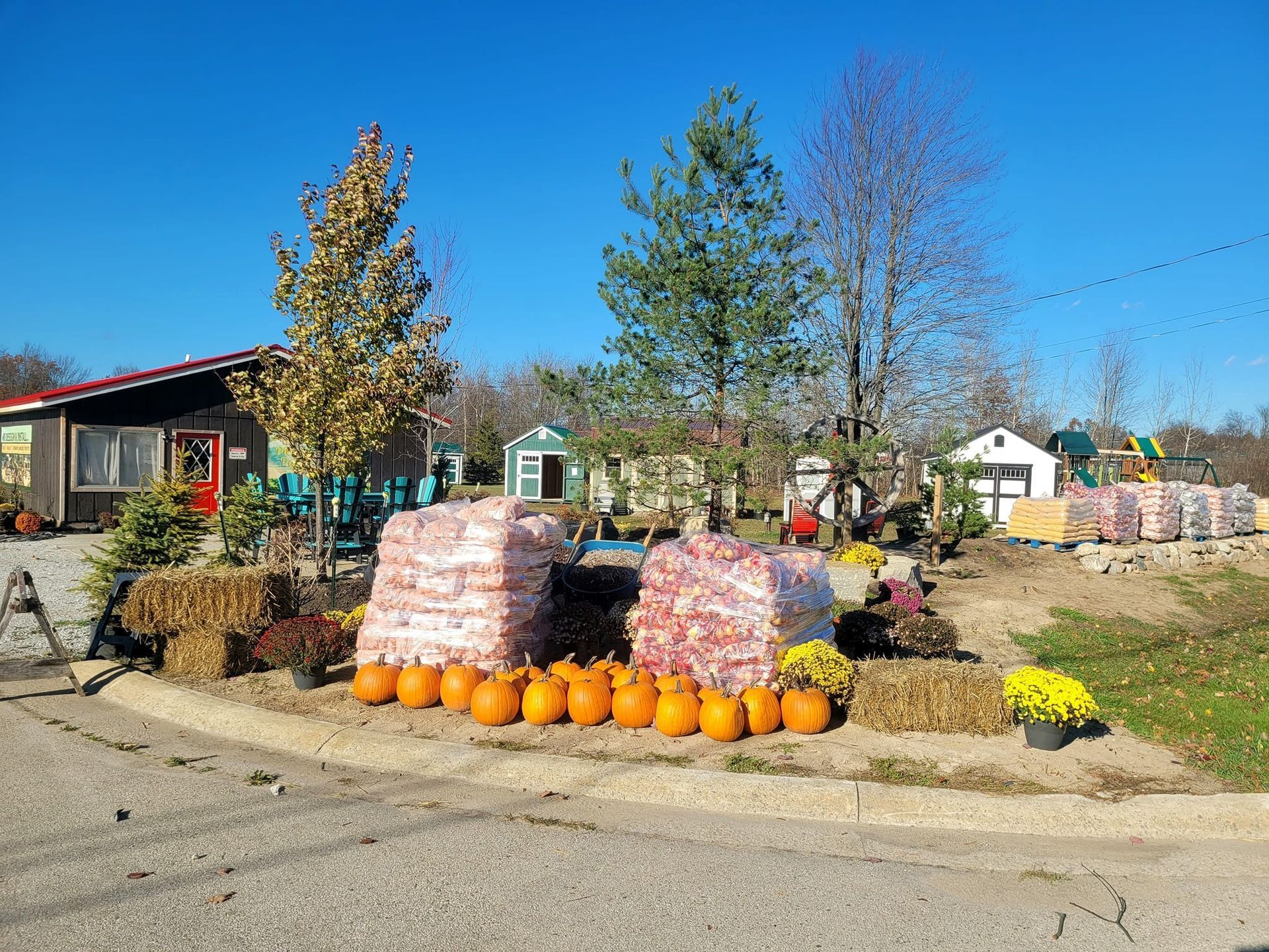 Autumn farm stand with hay bales, pumpkins, and mums. Bags of produce and small buildings in the background.