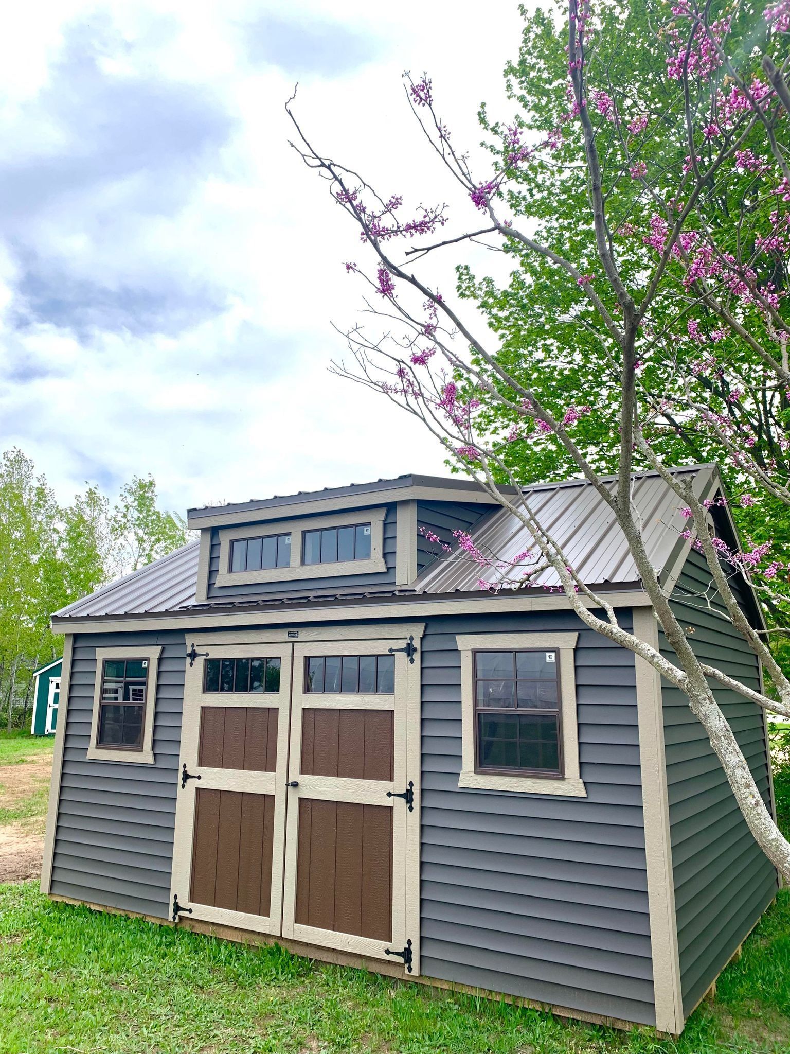 Gray shed with brown doors, windows, and metal roof, beside a flowering tree.