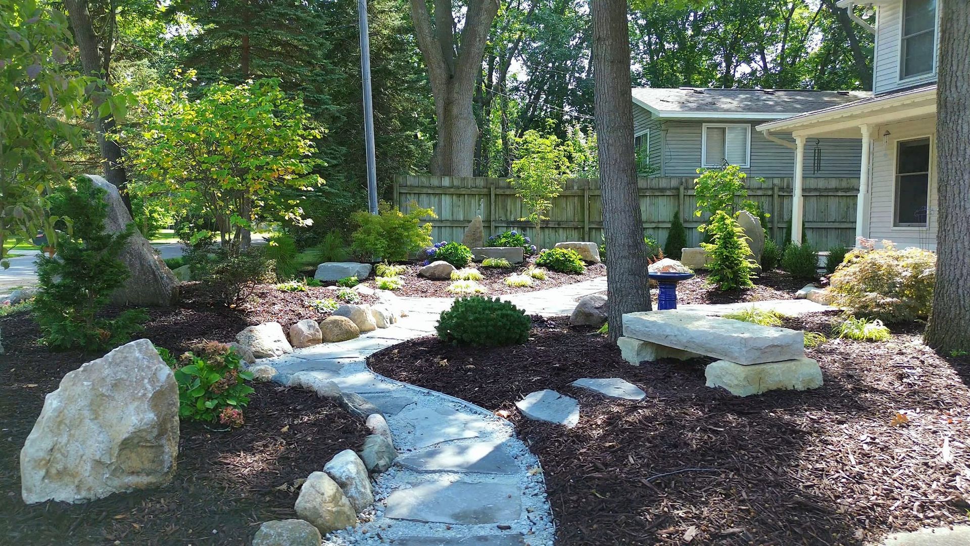 Stone path winds through a landscaped yard with trees, a bench, and a house.