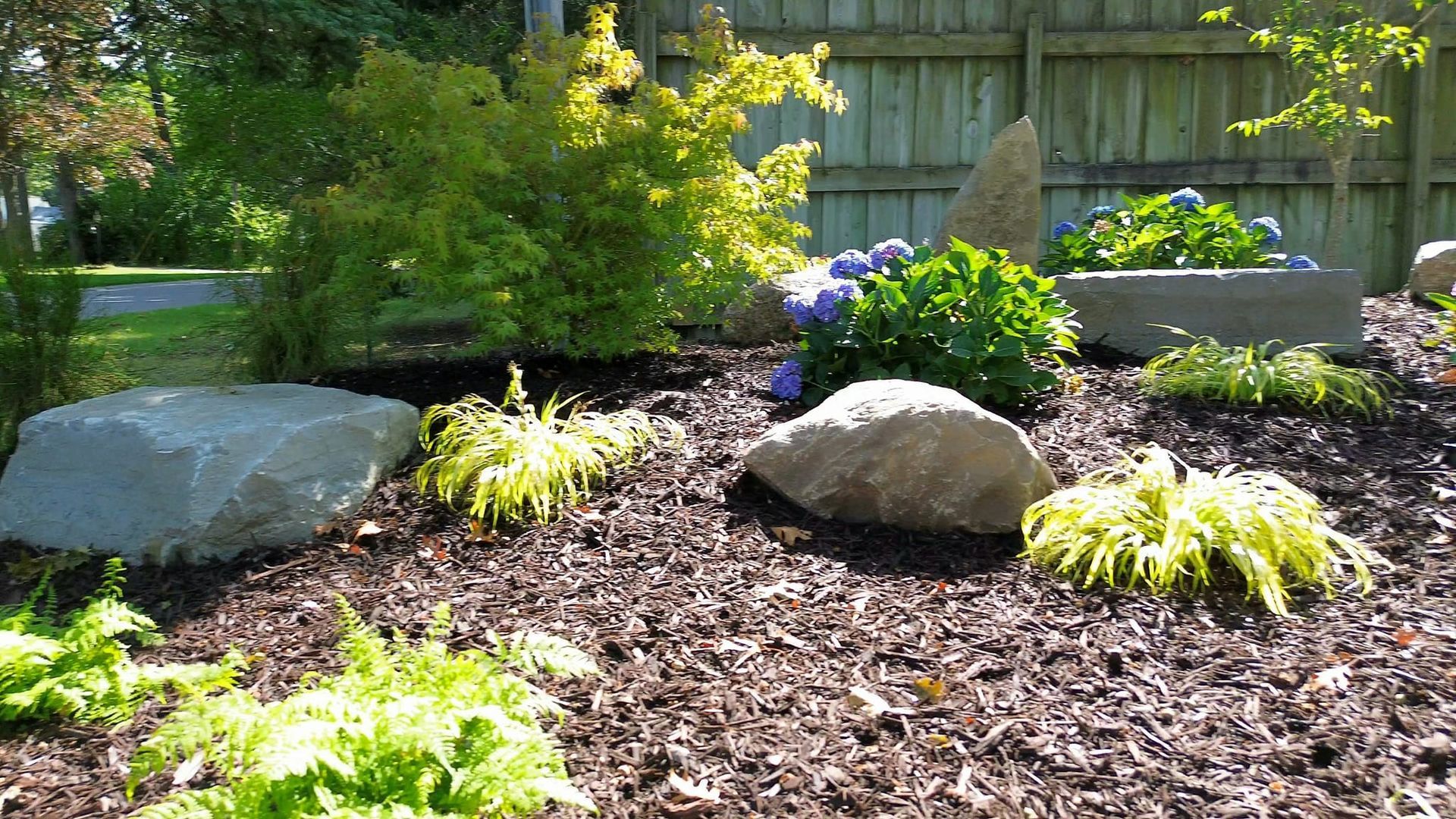 Landscaped garden bed with large rocks, mulch, and colorful plants; wooden fence background.
