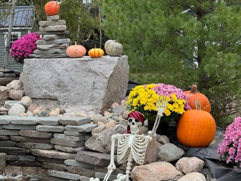 Halloween display with pumpkins, flowers, and a skeleton near a stone structure.