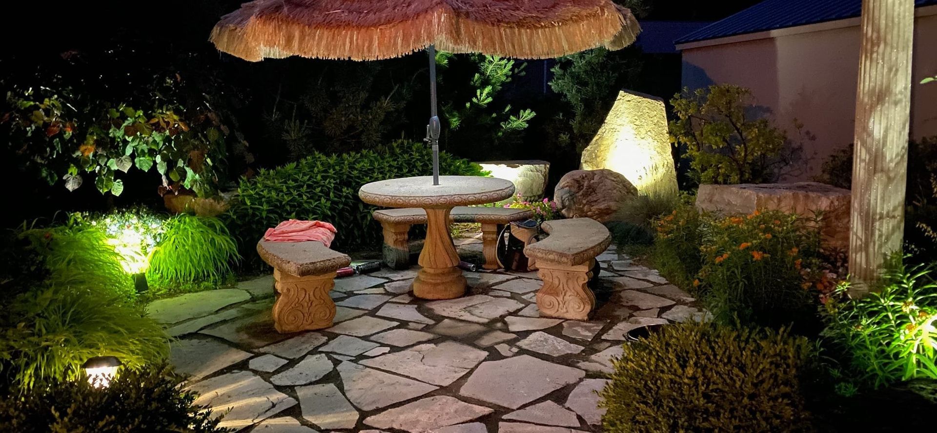A stone table and benches sit on a flagstone patio, under a fringed umbrella, illuminated by garden lights at night.