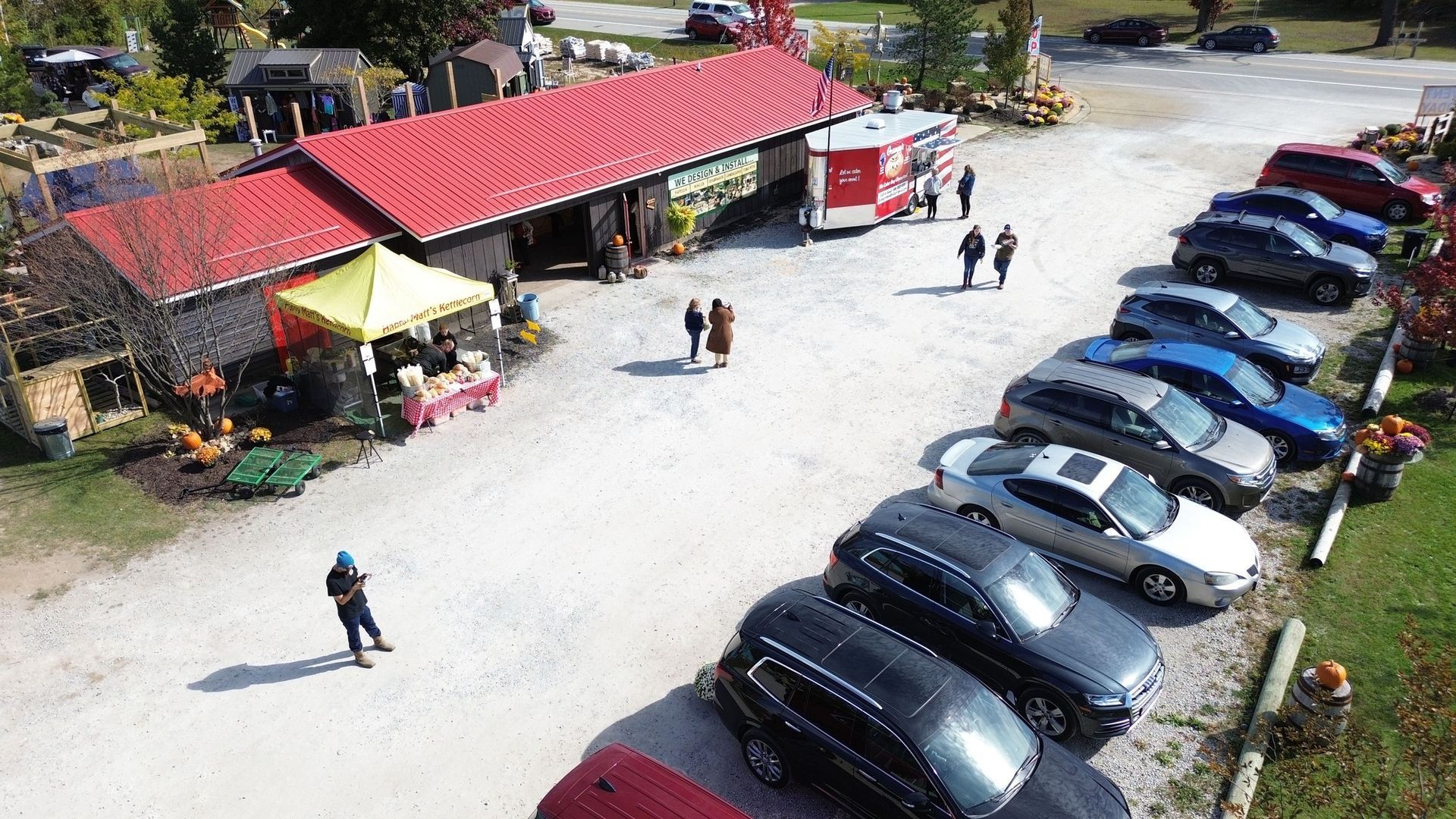 Aerial view of a farm stand with red roof, cars parked in front, people walking, and pumpkins.