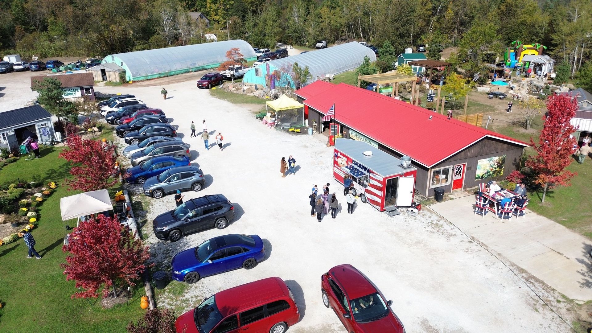 Aerial view of a farm with a red-roofed building, parked cars, and people gathered on a sunny day.