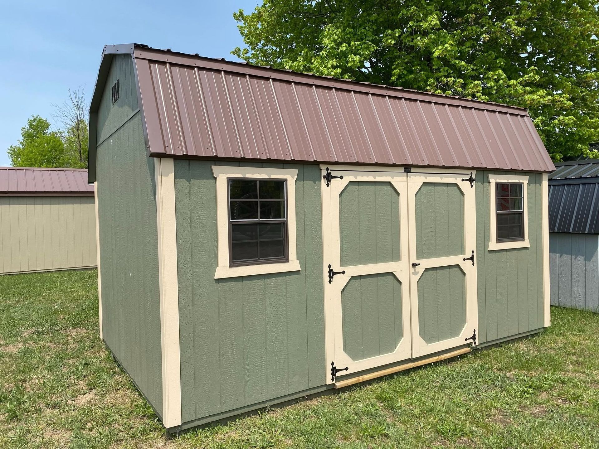 Green shed with brown metal roof, two windows, and double doors on a grassy lawn.