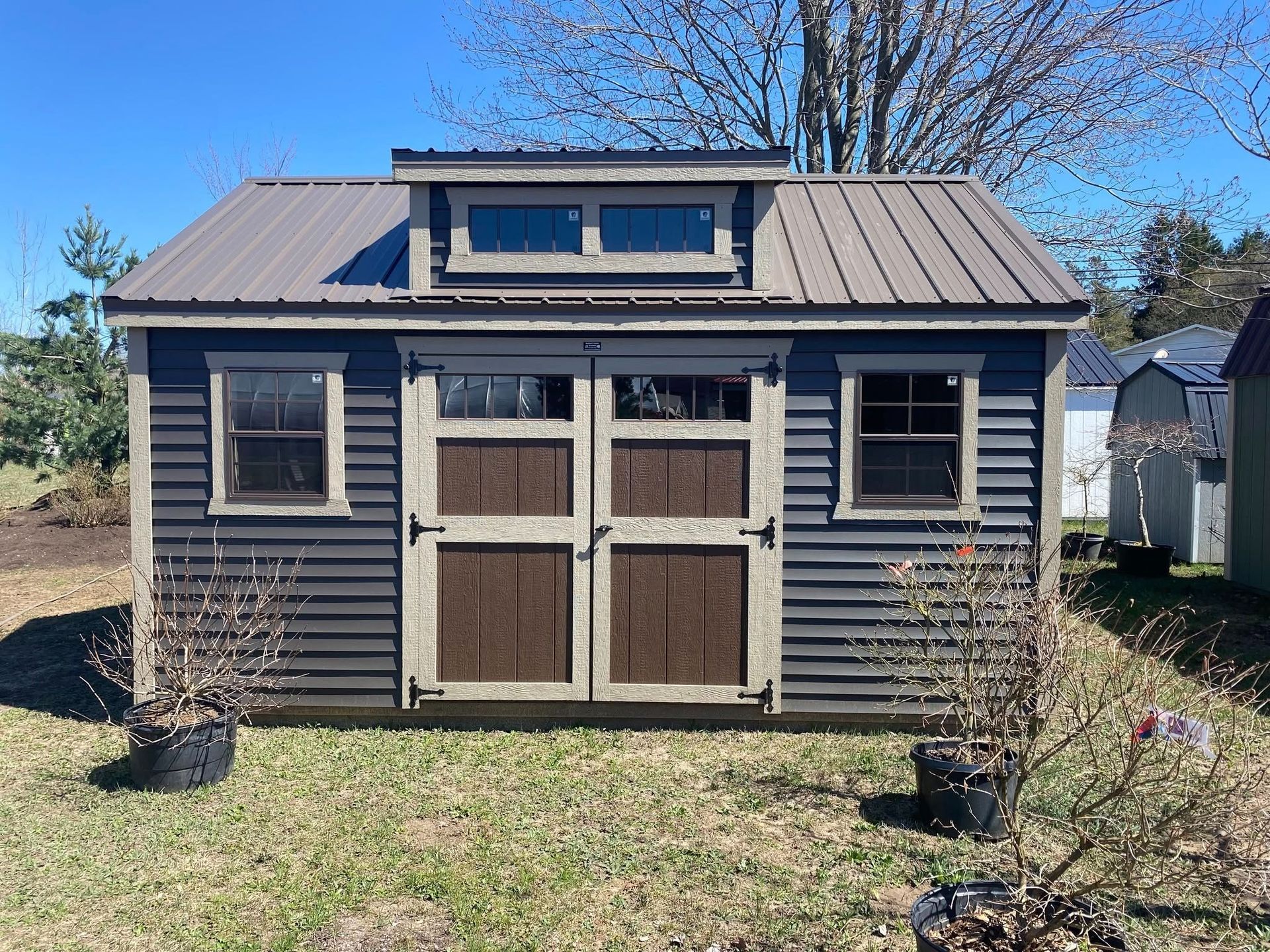 Blue shed with brown metal roof and double doors, two windows, and small windows above doors, sitting on green grass.
