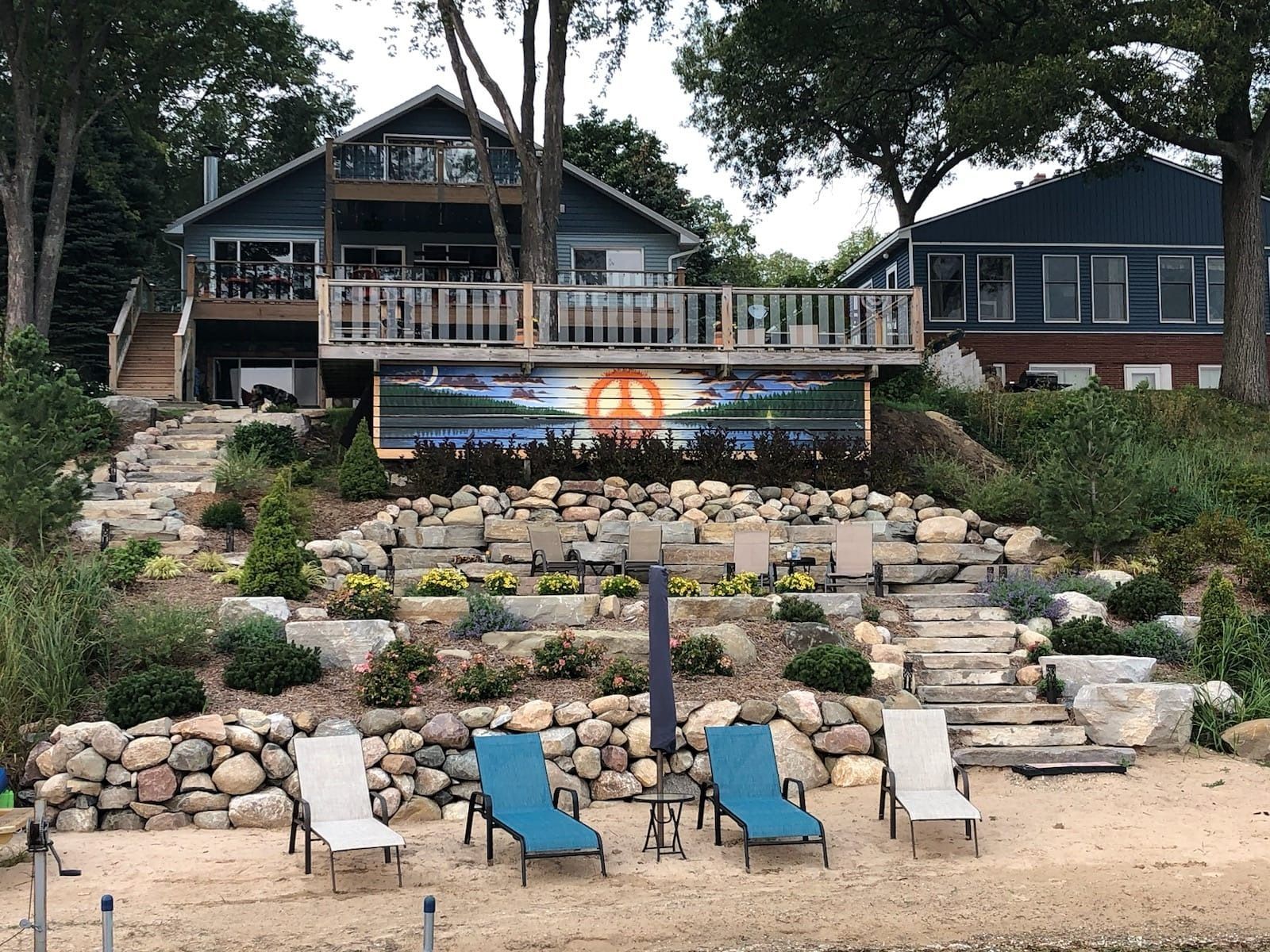Lakeside house with tiered landscaping and beach chairs. Teal and beige chairs on sand, blue house with wooden deck.