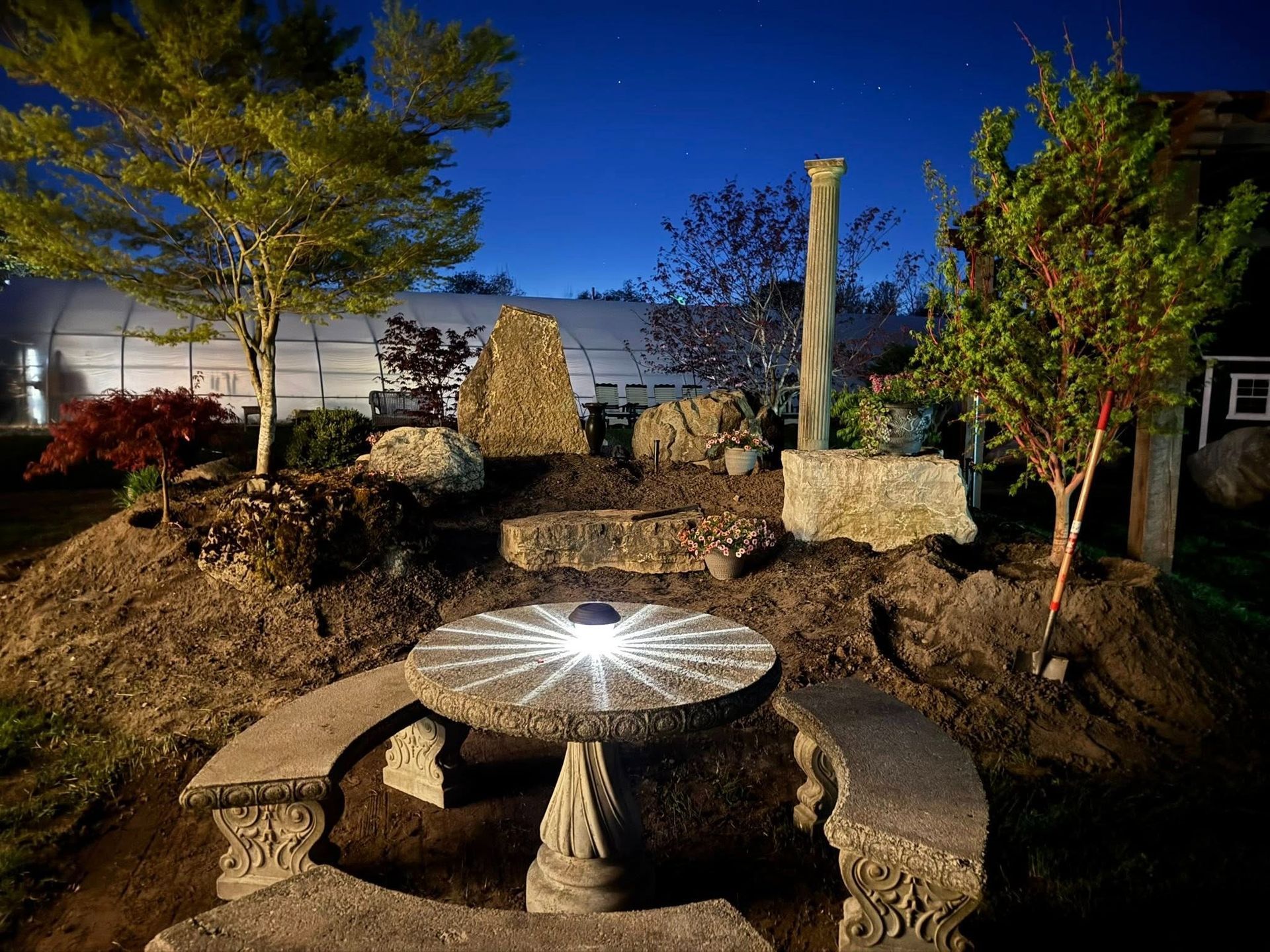 Stone garden setting with table, benches, and pillar, illuminated at dusk.