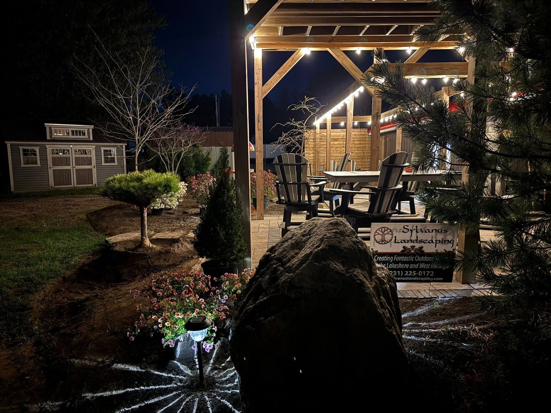 Nighttime backyard scene with a wooden pergola, string lights, and seating area. A shed is in the background.