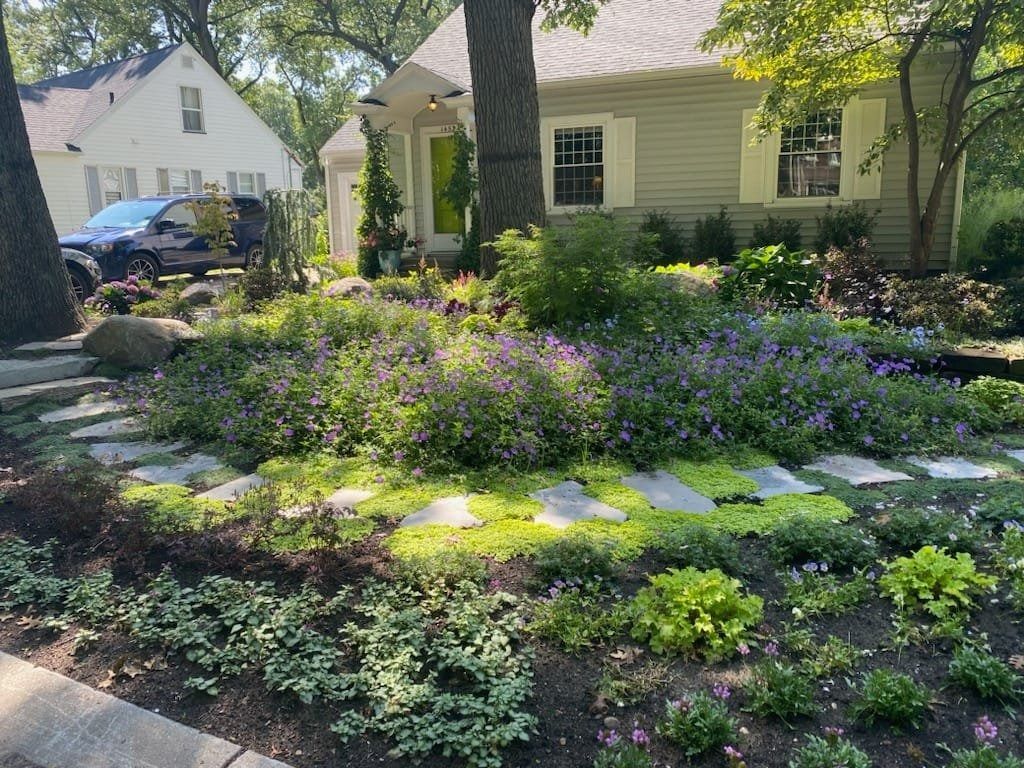 Landscaped front yard with flowering plants, stone path, and a light green house.