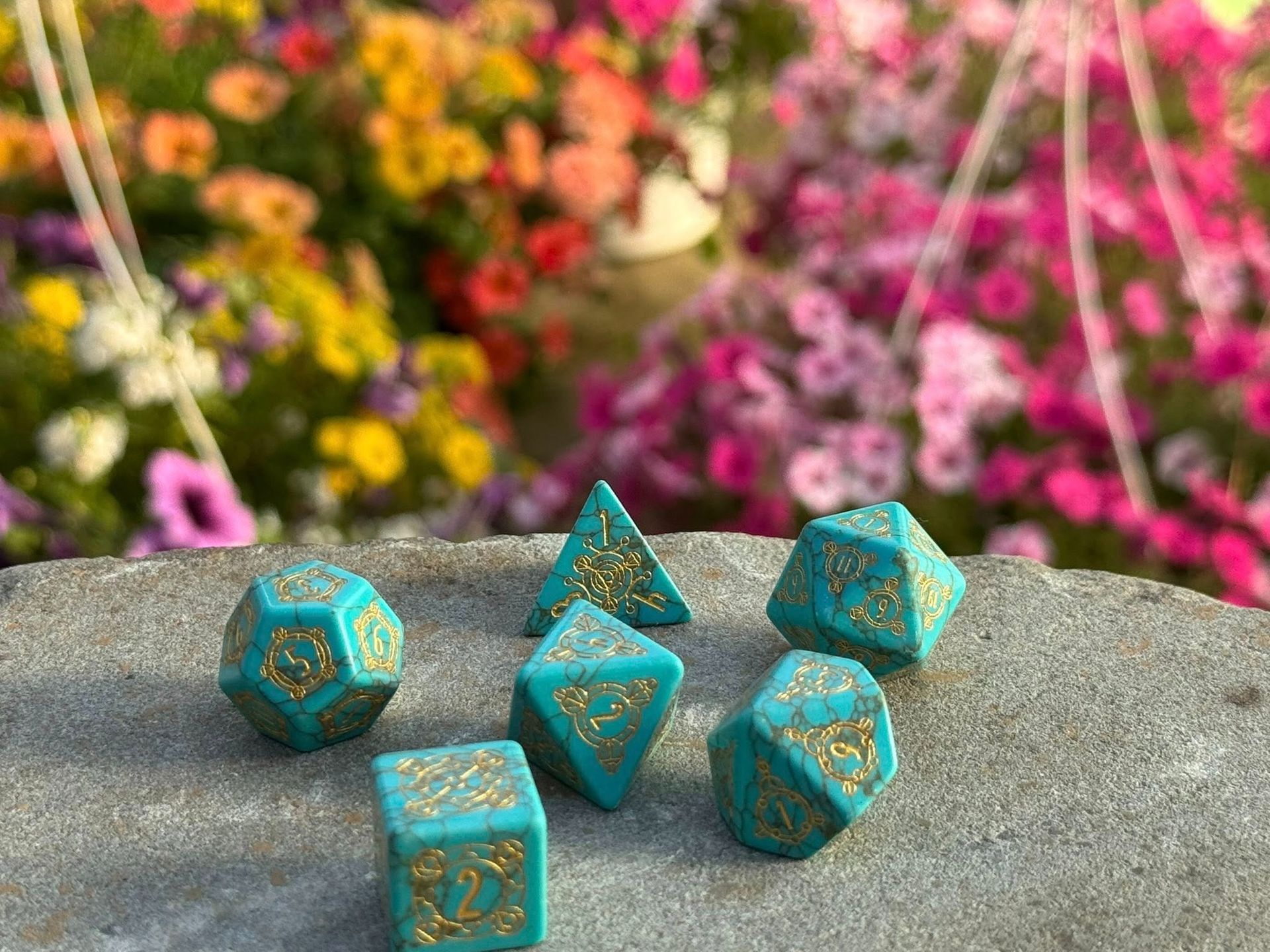 Set of teal dice with gold markings on a stone surface, blurred flowers in the background.