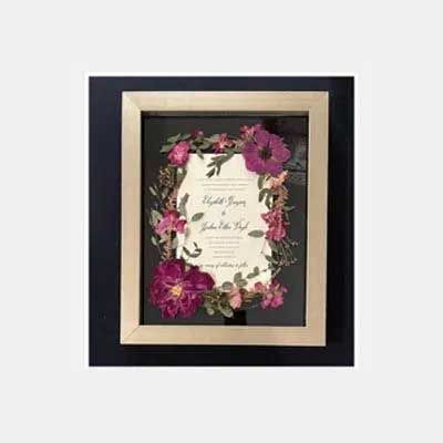 Framed wedding invitation with pink flowers, beige and black frame, on a light grey background.