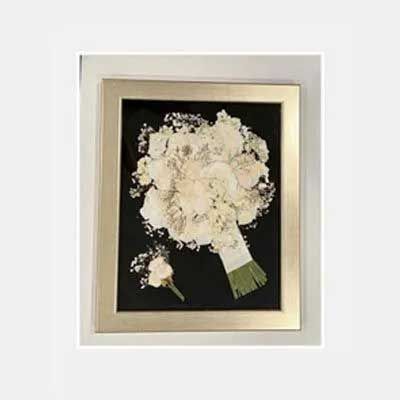 Framed wedding bouquet and boutonniere in gold frame on black background.
