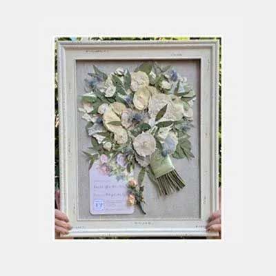 Framed pressed flower bouquet with a small tag, held by hands. Soft colors: cream, green, and blue.