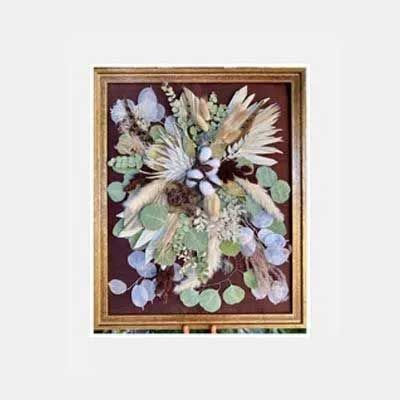 Dried floral arrangement in a gold-framed shadow box, featuring white, green, and brown foliage.