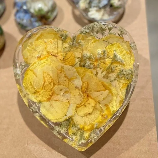 Heart-shaped resin paperweight with yellow petals and green inclusions, sitting on a brown surface.
